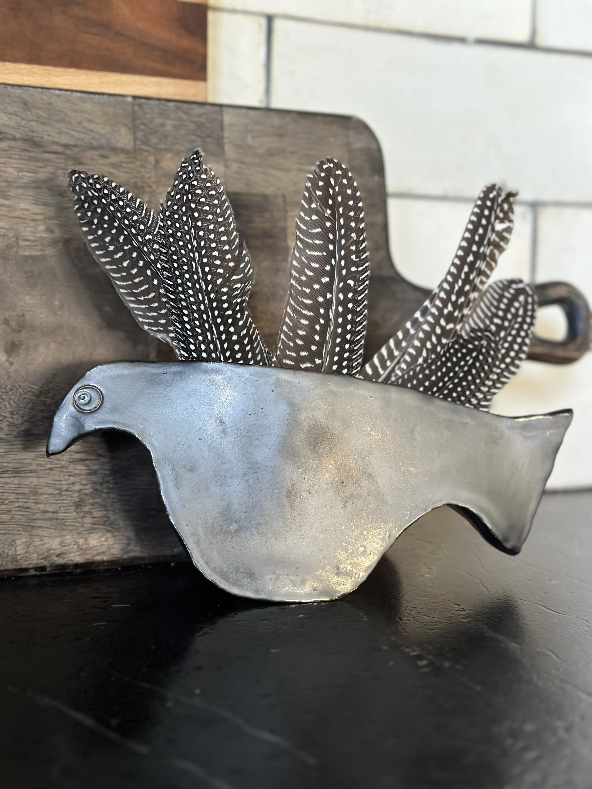 Decorative bowl shaped like a bird, filled with black and white patterned feathers, placed on a dark surface with a wooden and tiled background.