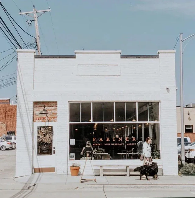 Front of a white brick bakery and wine merchant store named 'Fariner.' Large glass windows with people inside, a woman with a dog outside, and street utility poles nearby.