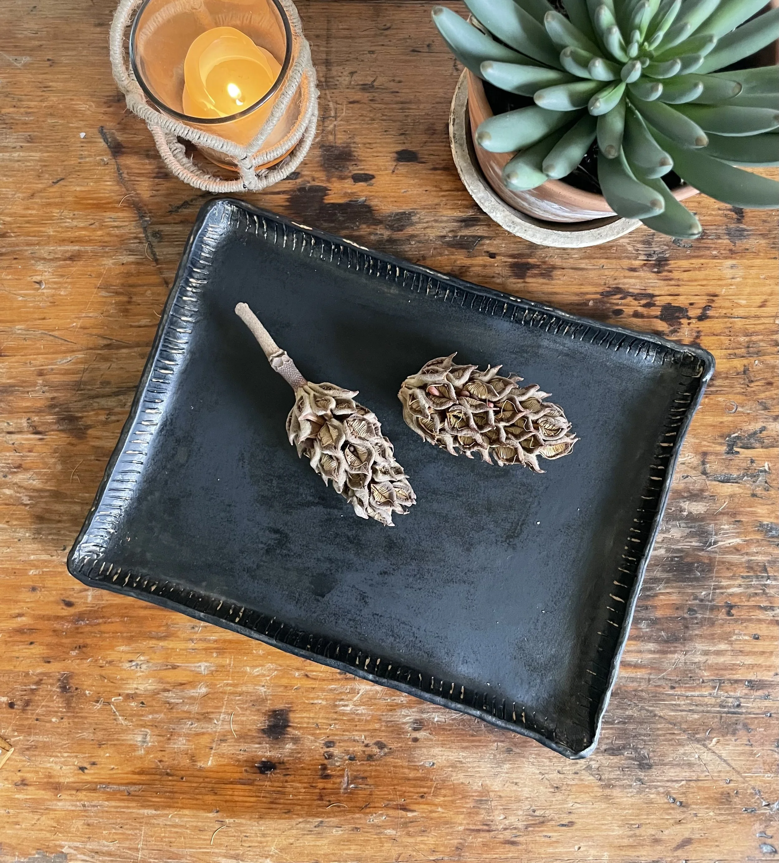 A black rectangular tray on a wooden table, holding two dried lavender buds with stems. To the top right, there is a green succulent plant in a terra cotta pot. To the top left, a lit candle in a jar wrapped with a decorative string, placed on a smal