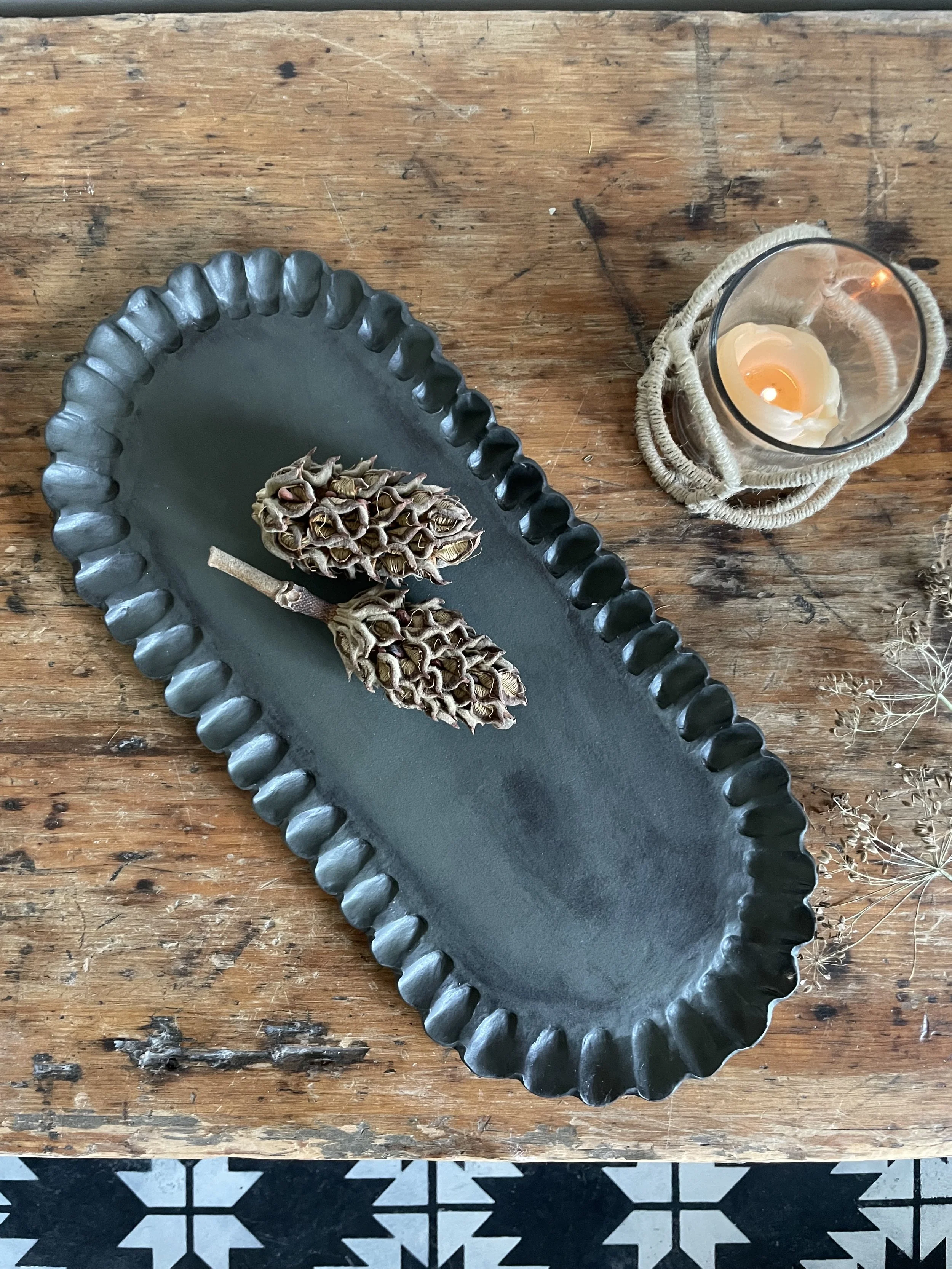 A black ceramic platter with two dried seed pods on it, placed on a rustic wooden table. To the right, there is a glass candle holder with a lit candle wrapped in a decorative rope.