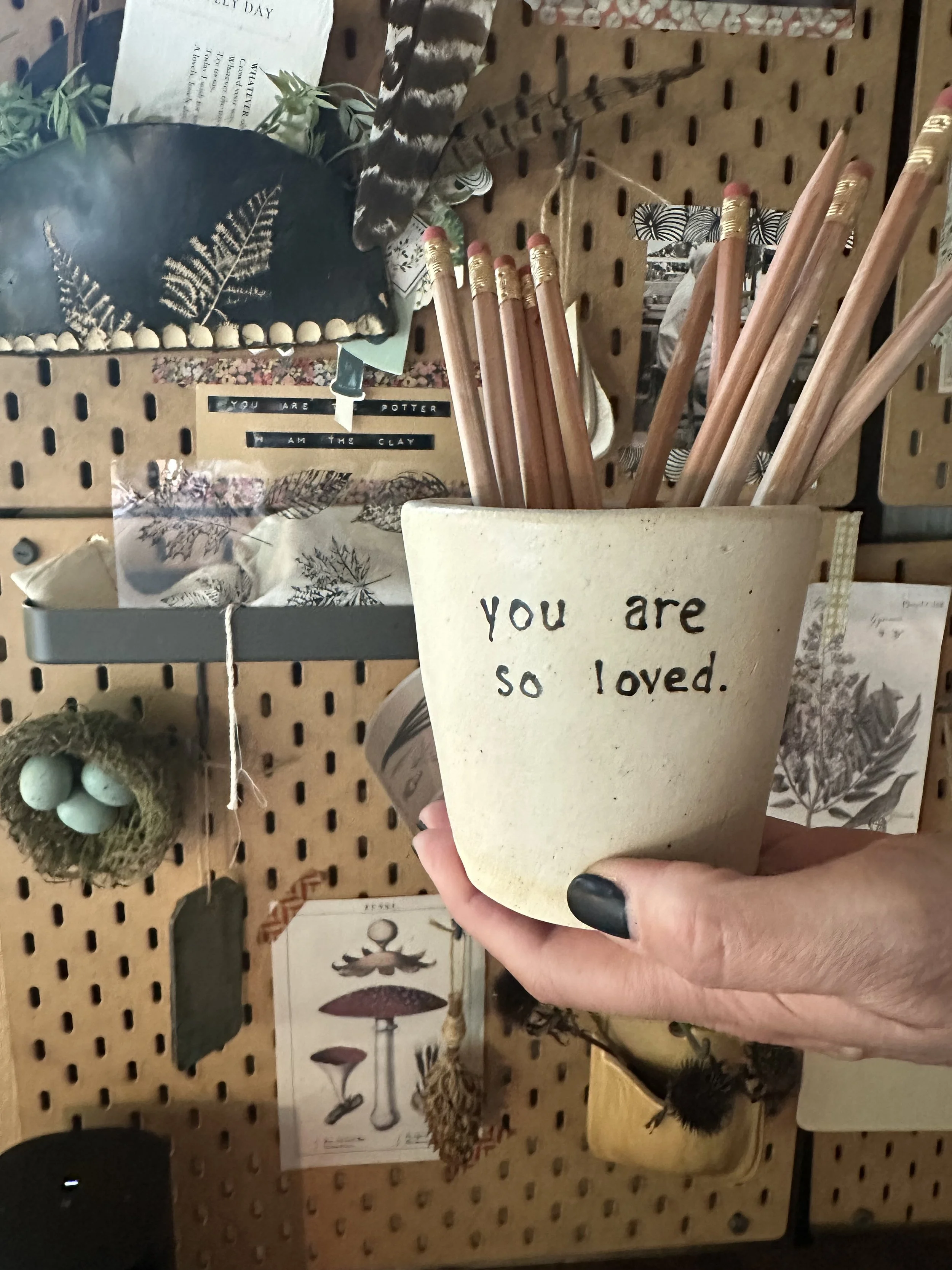 A hand holds a beige paper cup with the words "you are so loved" written on it, containing pencils with pink erasers. The background shows a pegboard with various decorative items, including a small nest with eggs, pictures, and other decorations.