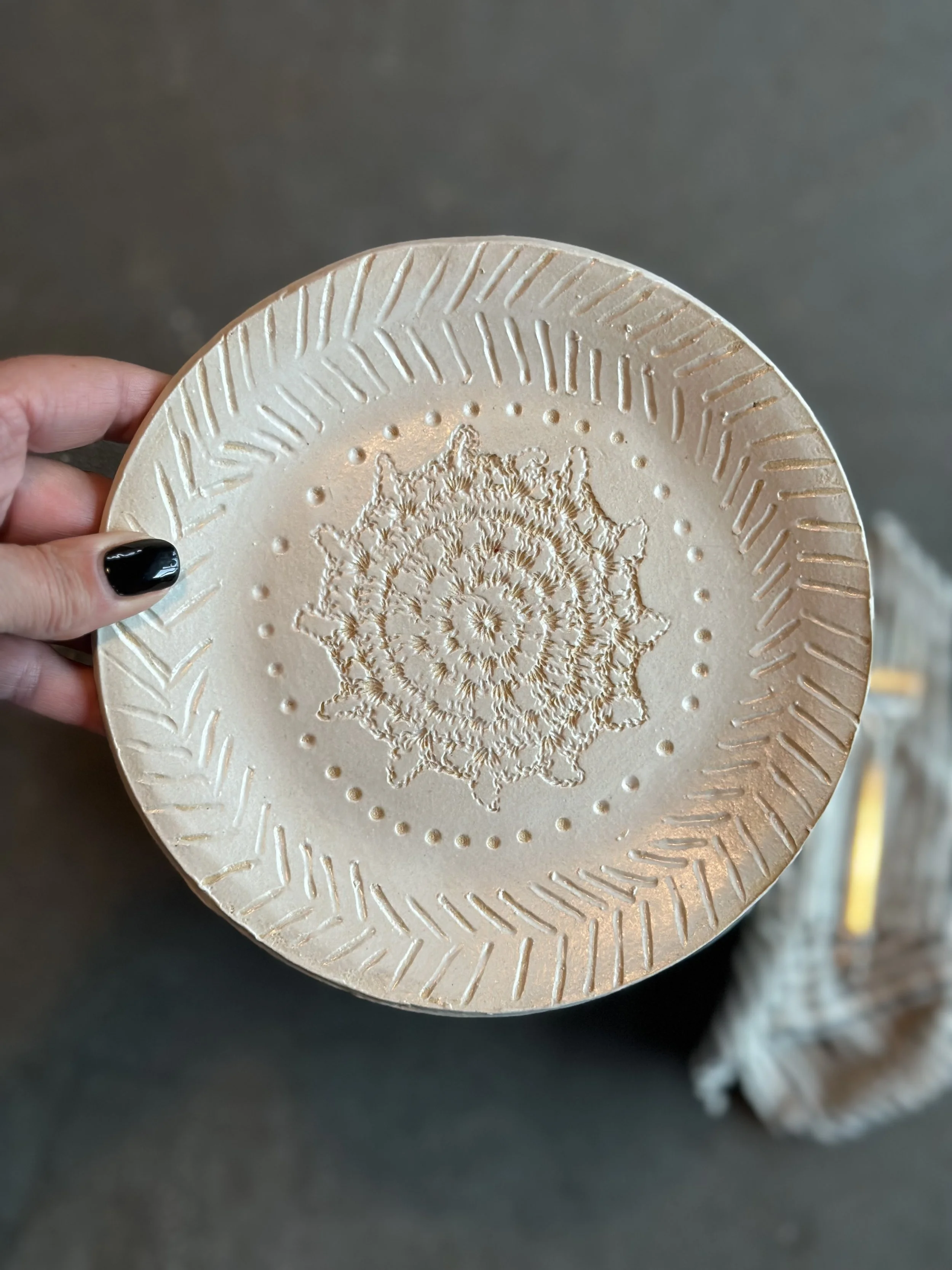 Beige paper plate with a decorative embossed pattern in the center and a geometric border, held by a hand with black nail polish.