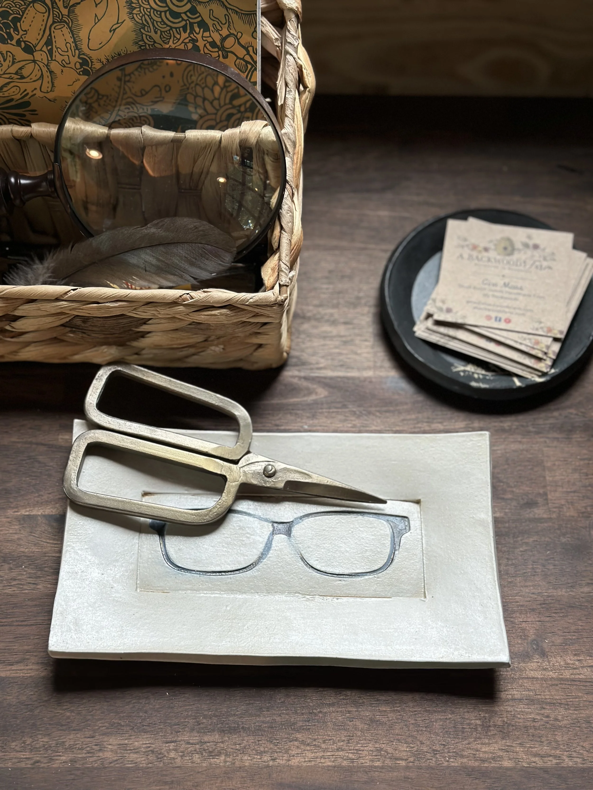 A box with a mirror and a feather inside, a black tray with stacked business cards, a pair of glasses on a box, and a large pair of metal scissors on a wooden surface.