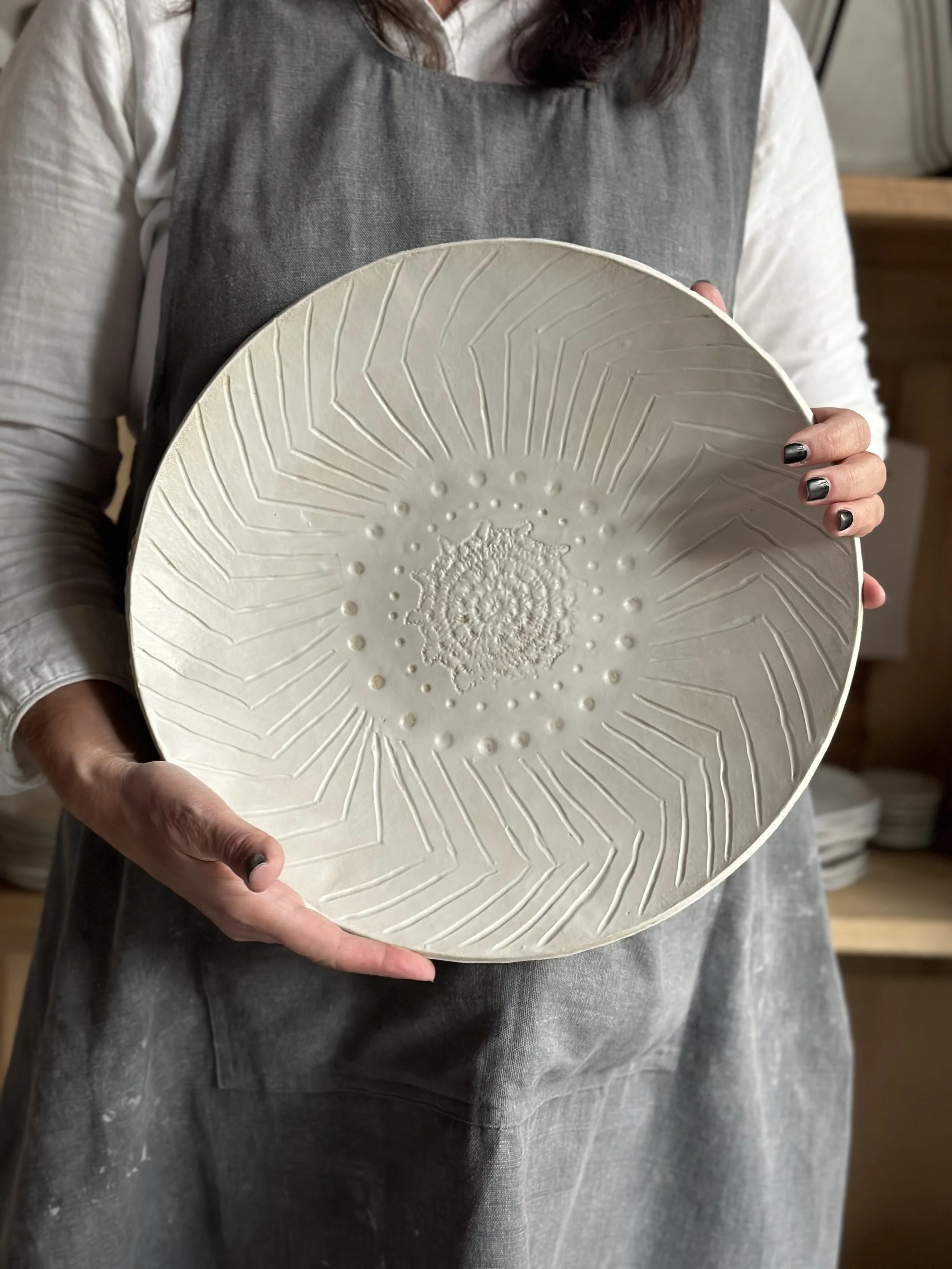 Person holding a large, round, textured ceramic plate with geometric and organic patterns, wearing a gray apron over a white long-sleeve shirt.