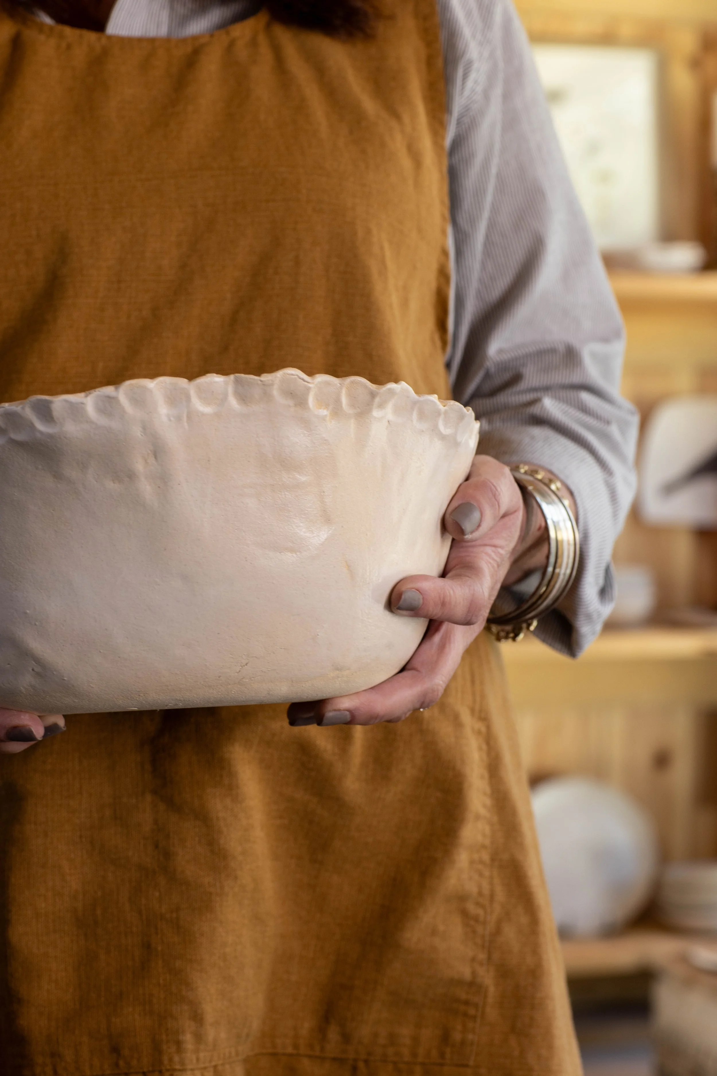 Person holding a large, unfinished ceramic bowl in a pottery studio, dressed in a brown apron and a light gray shirt.