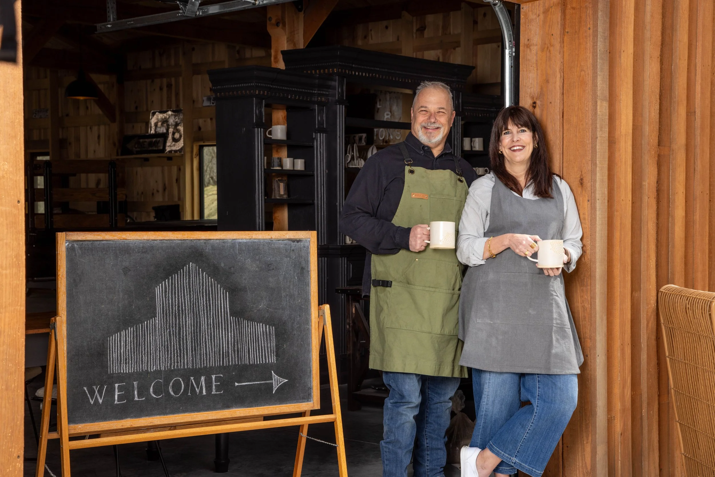 A smiling man and woman, both holding mugs, standing inside a cafe with wooden walls. A chalkboard sign with a house and the word 'Welcome' with an arrow is in front of them.