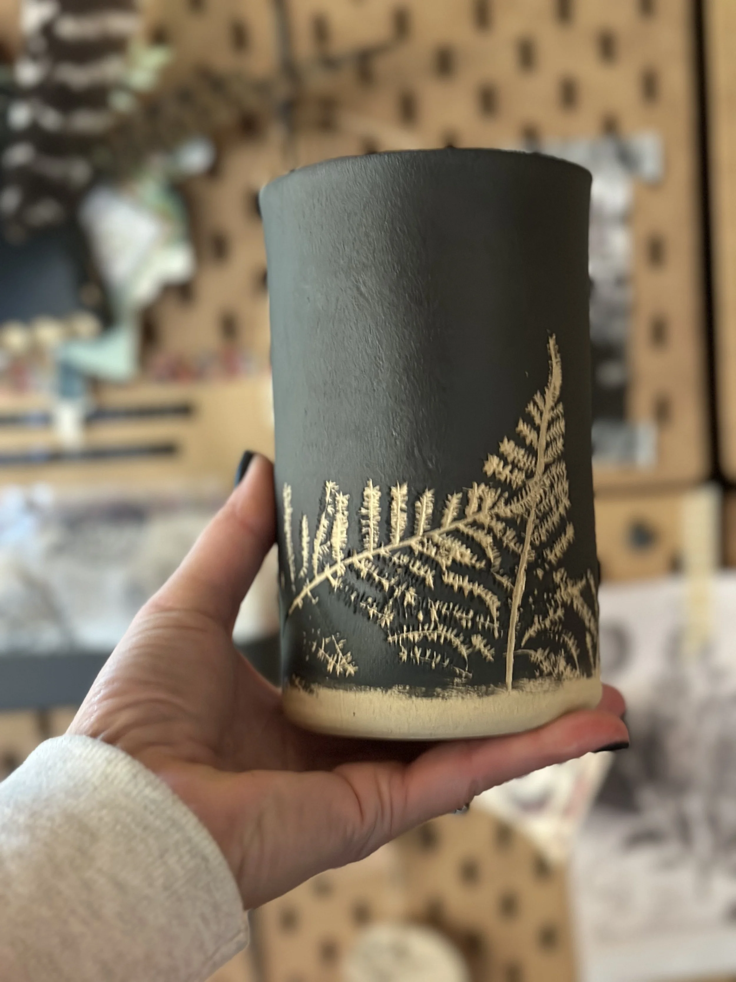 A person holding a black ceramic cup with white fern leaf designs, with a blurred background of a wooden pegboard display.