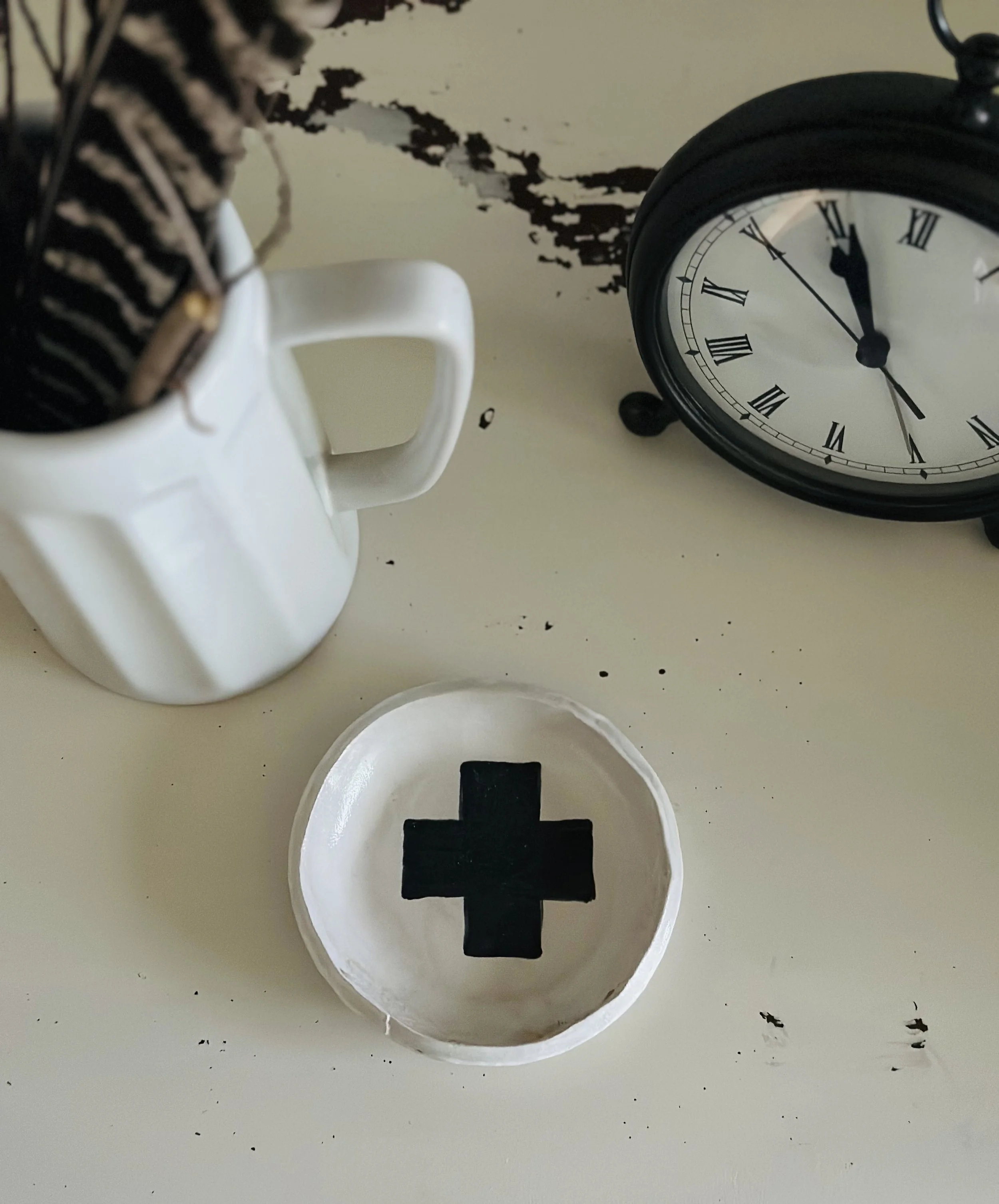 A white mug holding a potted plant with striped leaves, a black vintage clock showing 11:55, and a small white dish with a black cross symbol on it, all on a white surface with black markings.