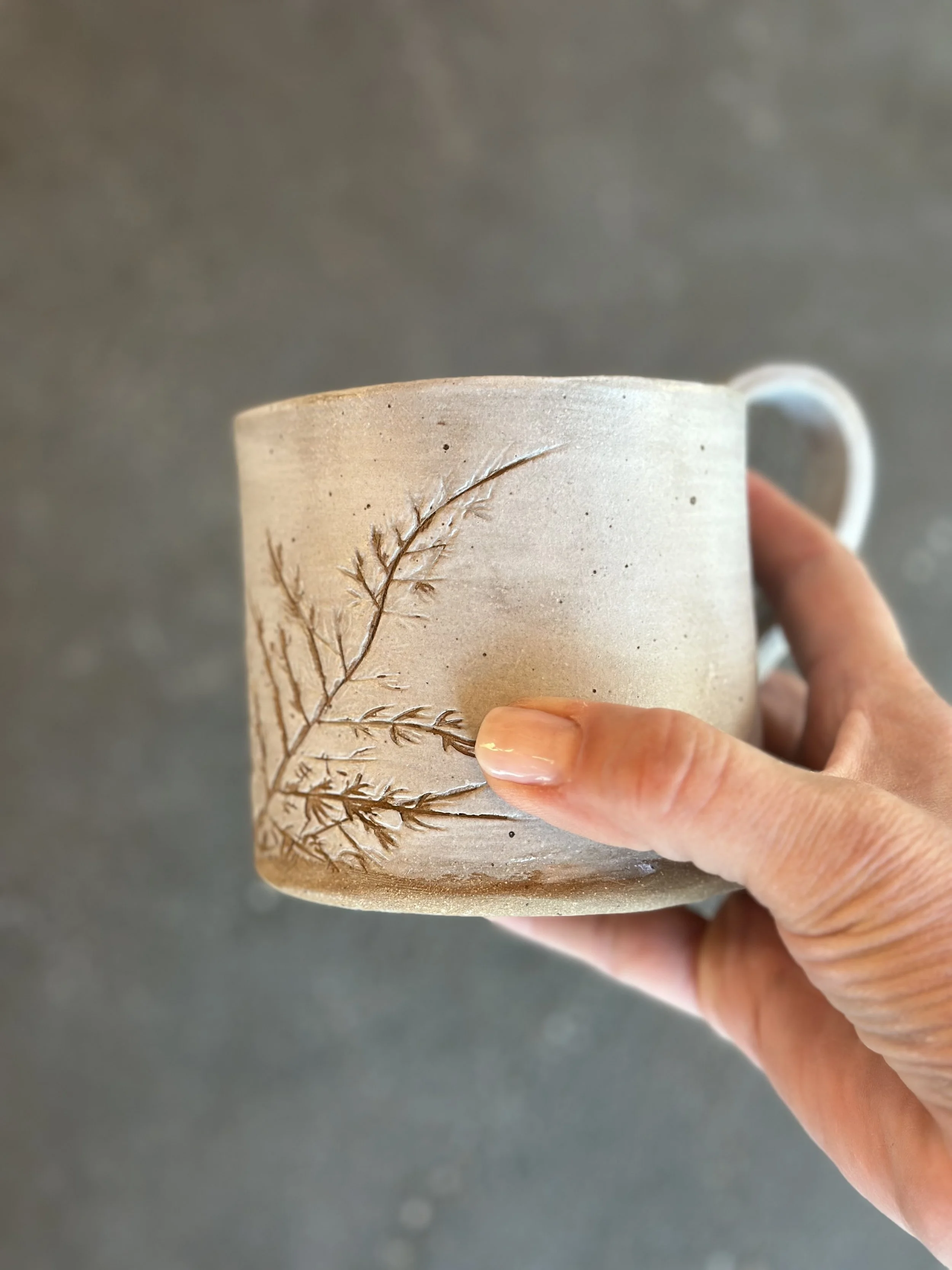 Hand holding a beige ceramic mug with a carved branch design.