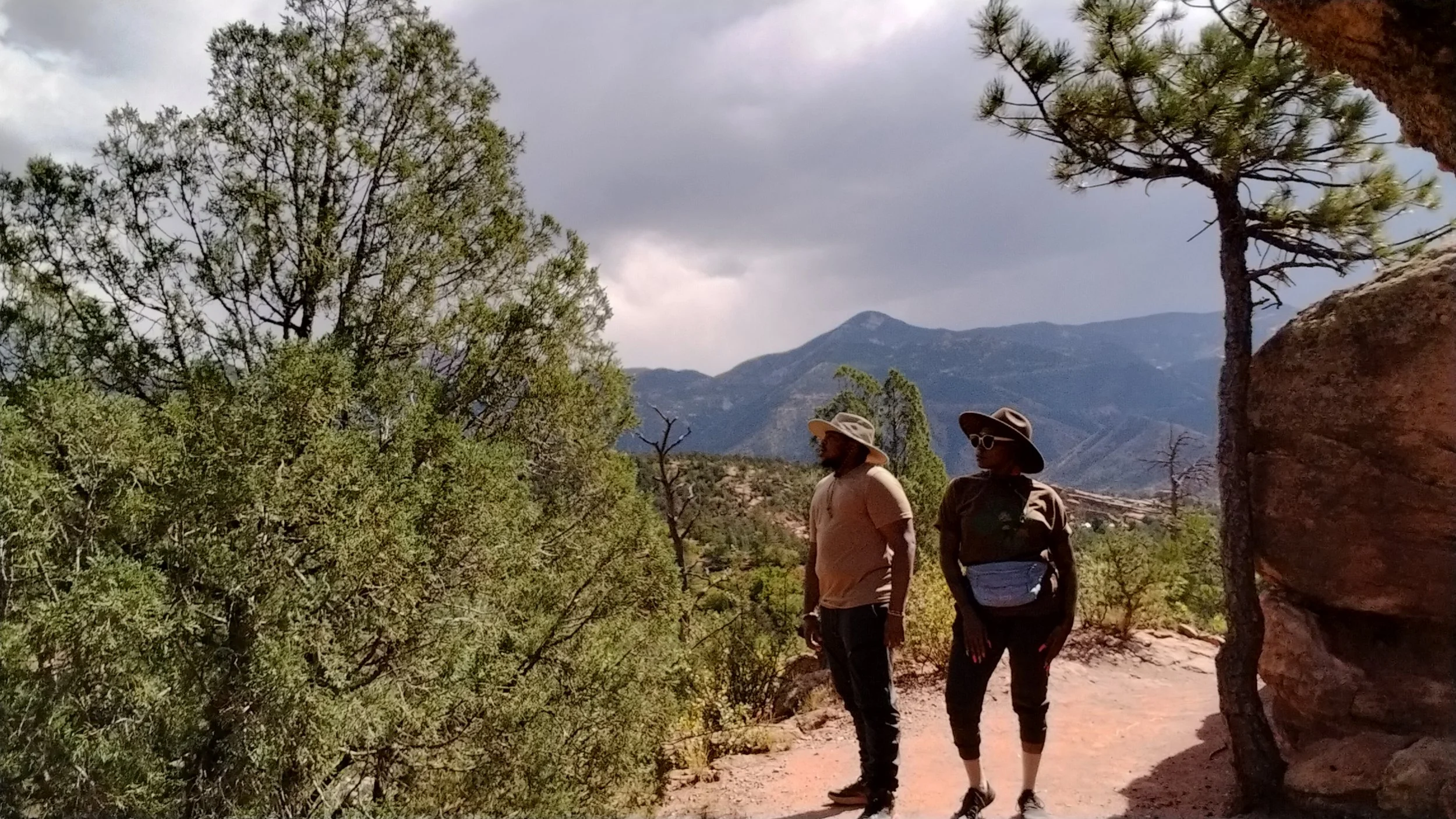 Two people hiking on a trail surrounded by trees and mountains in the background, wearing hats and sunglasses.