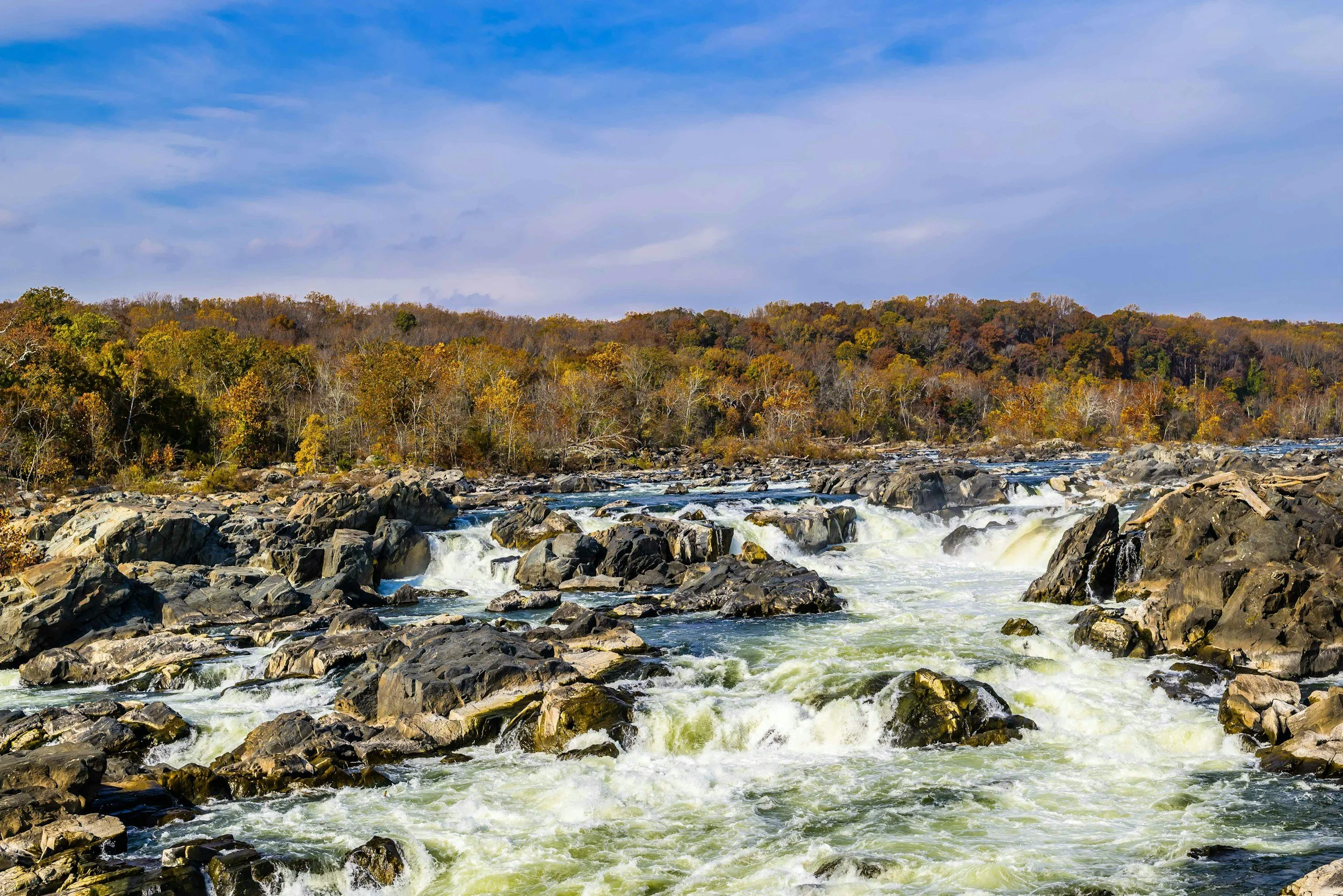 A river with rushing water flowing over rocks, with trees in fall colors and a partly cloudy sky in the background.