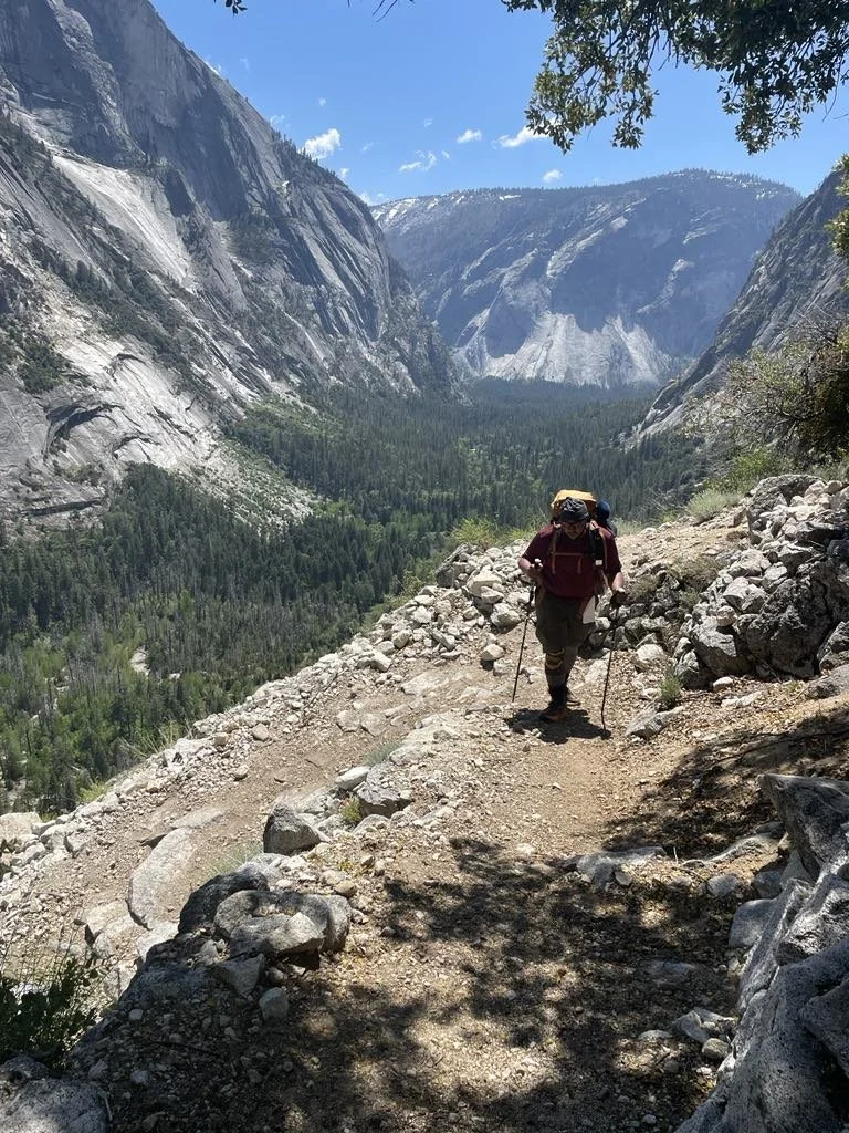 Hiker walking on a rocky mountain trail with a valley of dense green trees below and tall granite cliffs on either side, under a blue sky.