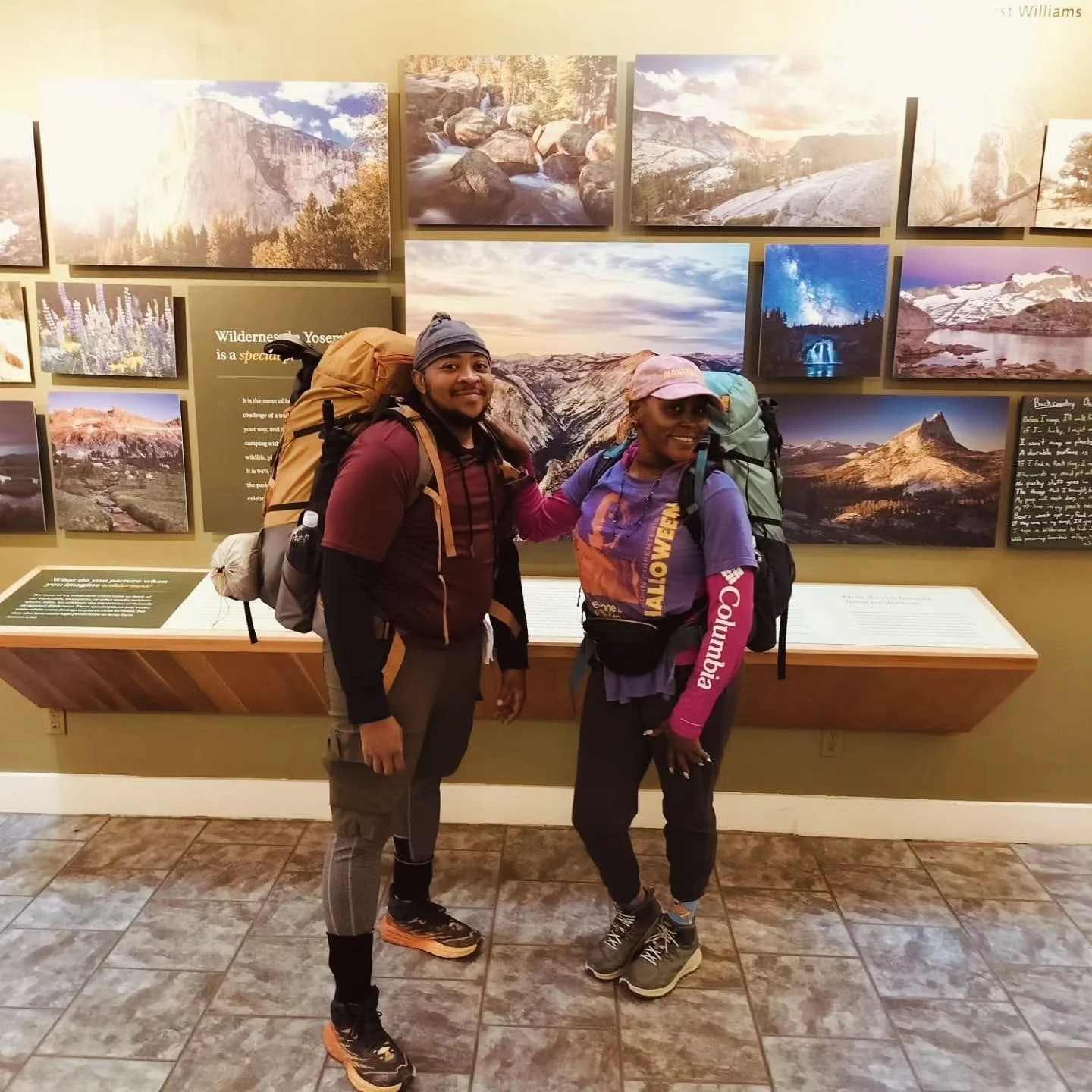 Two hikers with backpacks standing together inside a museum, in front of a wall display of landscape photographs of mountains, rivers, and stars.