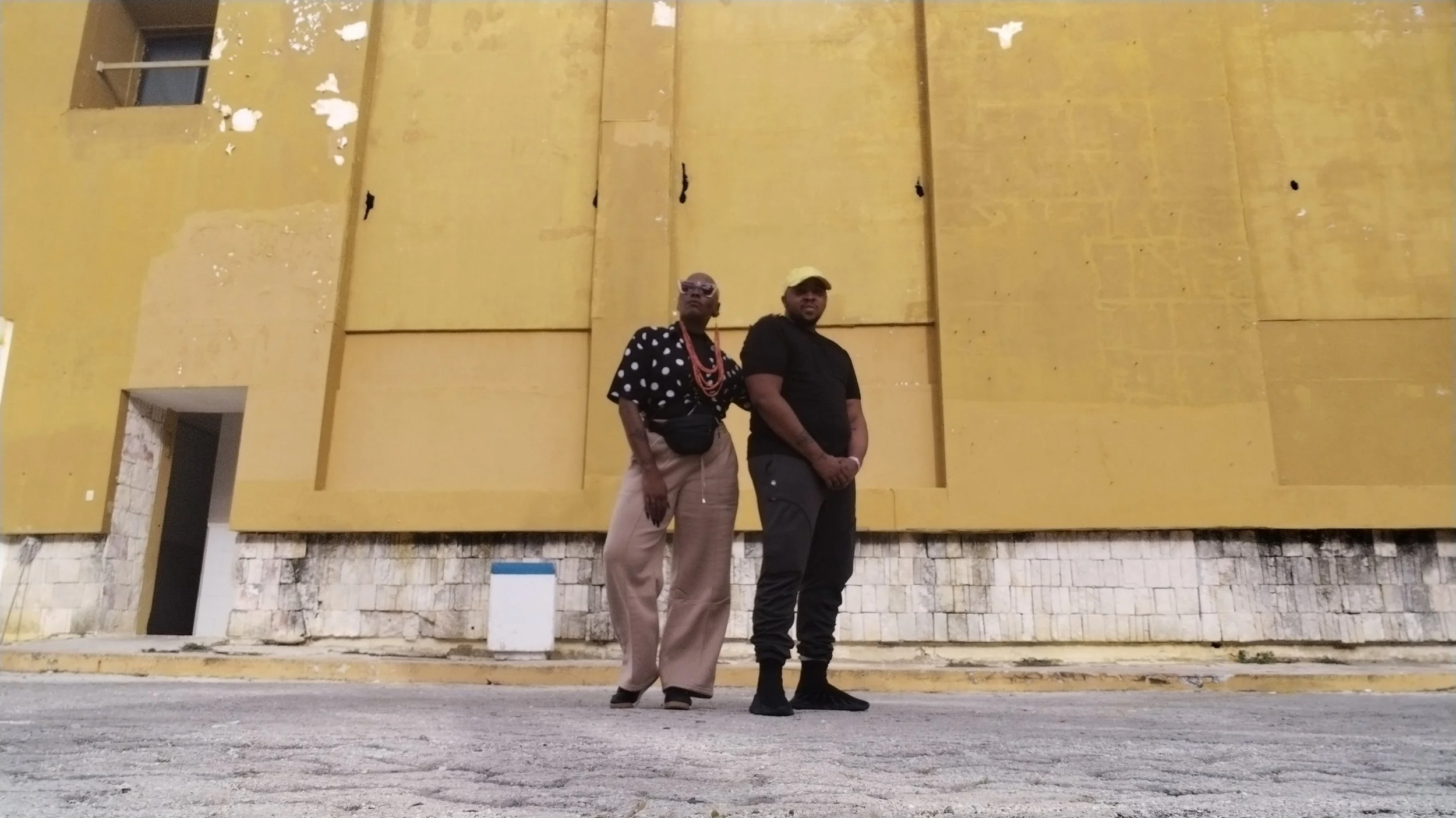 Two people standing on a street in front of a yellow building with a textured wall.
