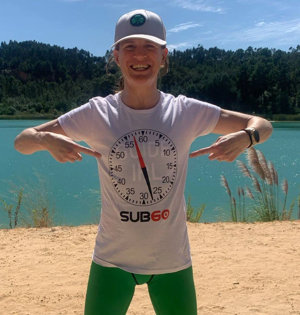 Woman smiling outdoors near a lake, wearing a white cap, white T-shirt, and green leggings, pointing to a large clock graphic on her T-shirt showing 25 minutes, with a background of blue water, trees, and a sandy shore.
