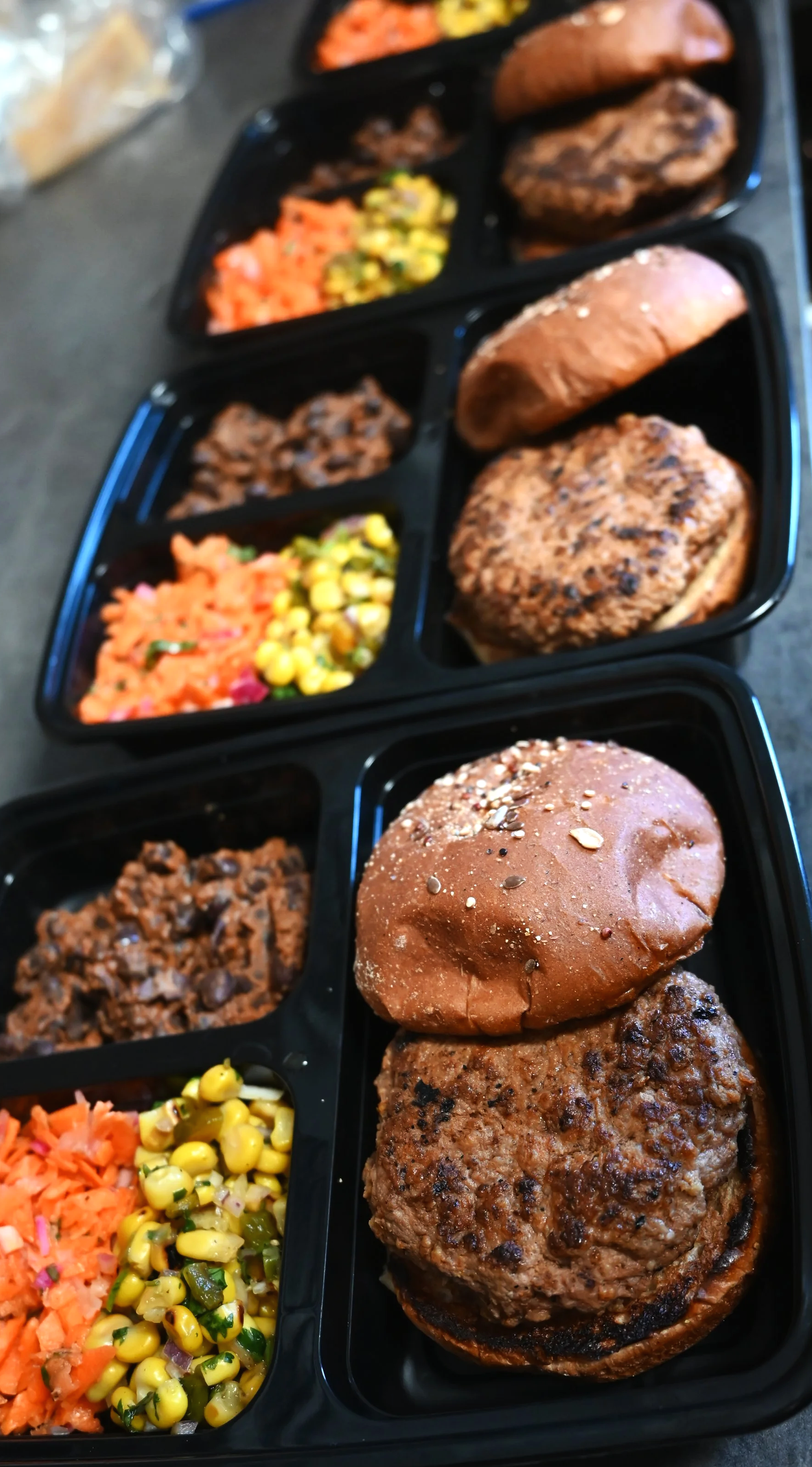 Pre-packaged meals with grilled burgers, buns, and assorted side dishes including corn salad, shredded carrots, and beans.