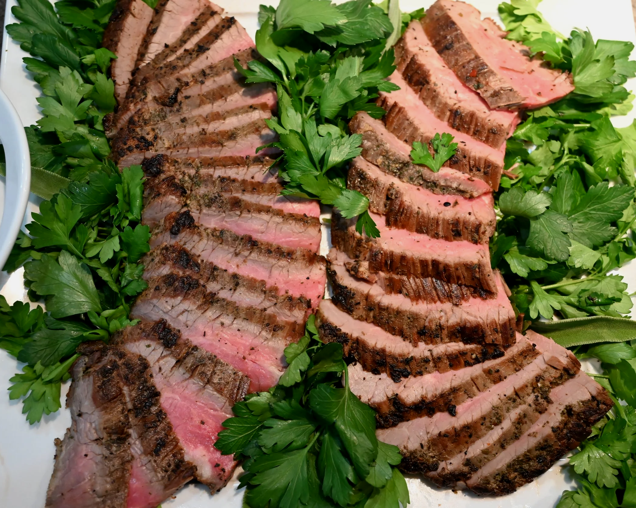 Sliced grilled steak with a pink center, garnished with fresh parsley and leafy greens, on a white platter.