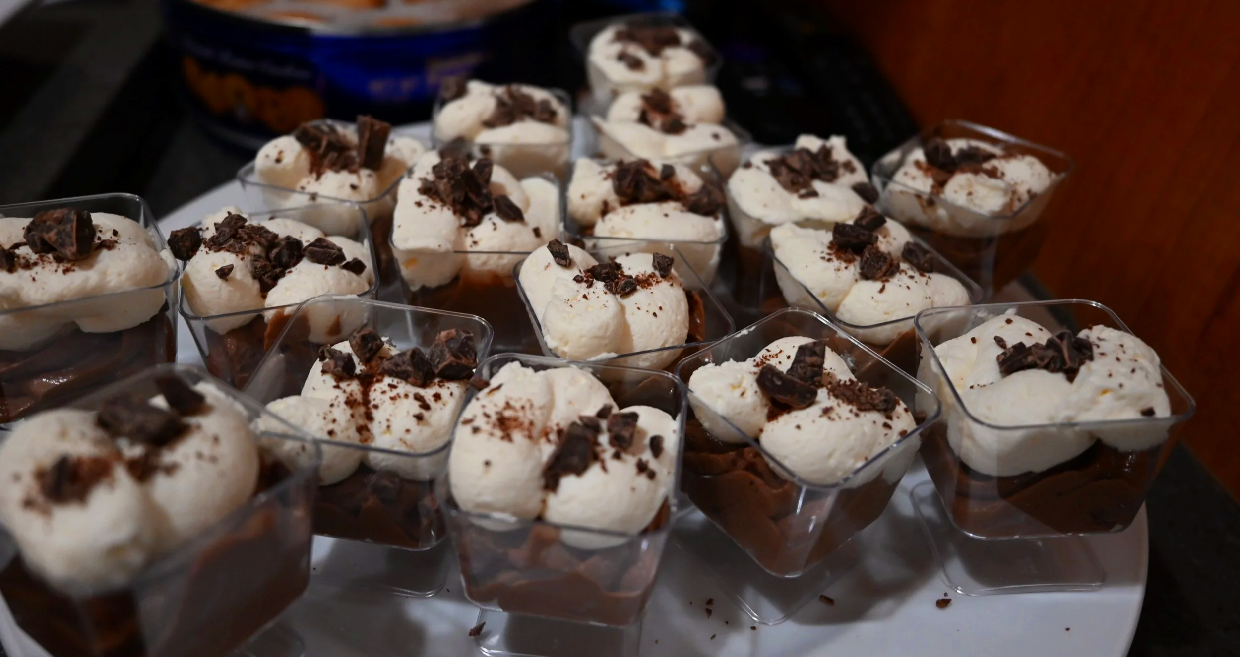 Chocolate dessert mousse cups topped with whipped cream and chocolate shavings on a white serving platter.