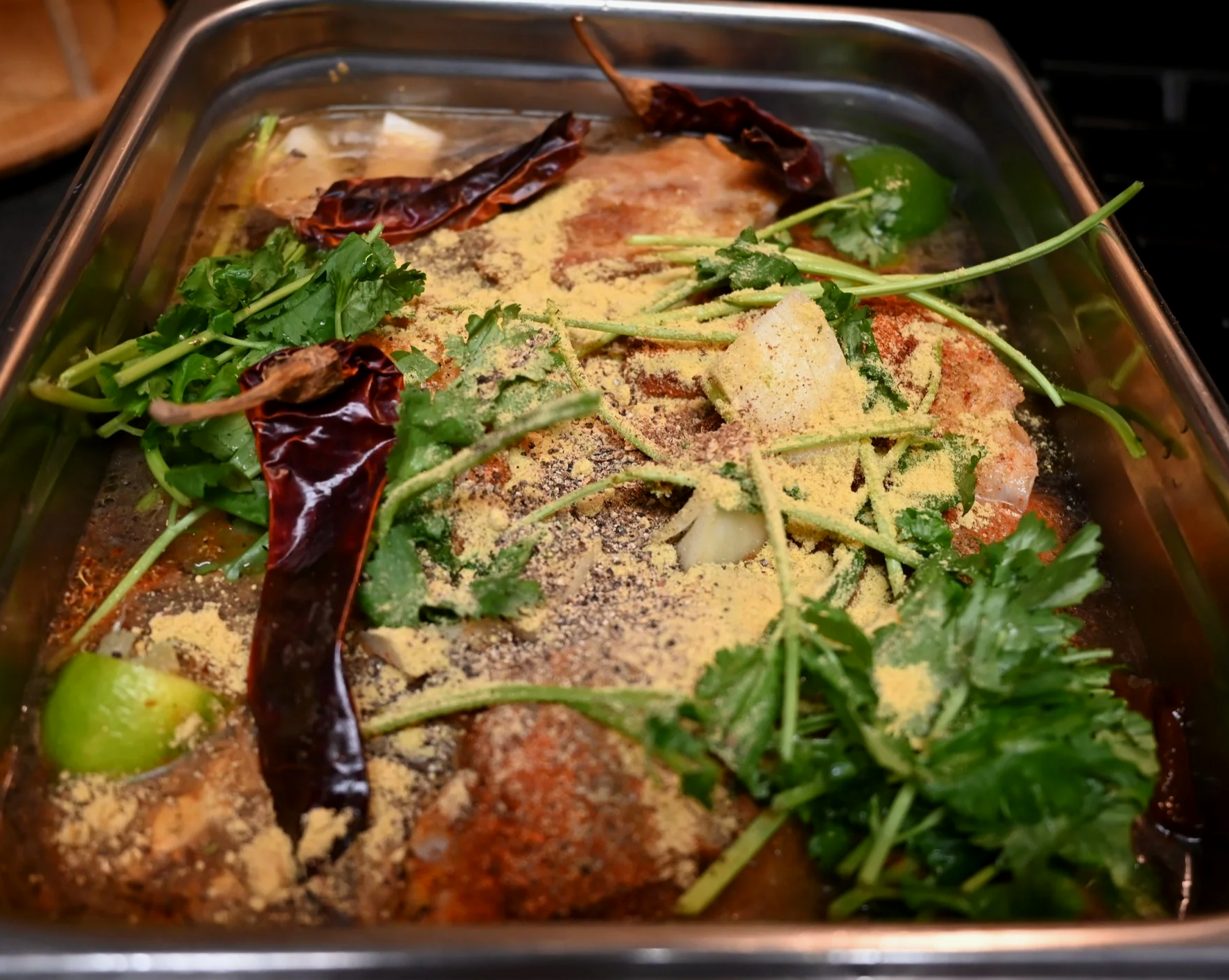 A aluminum pan containing seasoned raw chicken pieces topped with dried chili peppers, chopped cilantro, and sprinkled with seasonings ready to be braised in the oven.