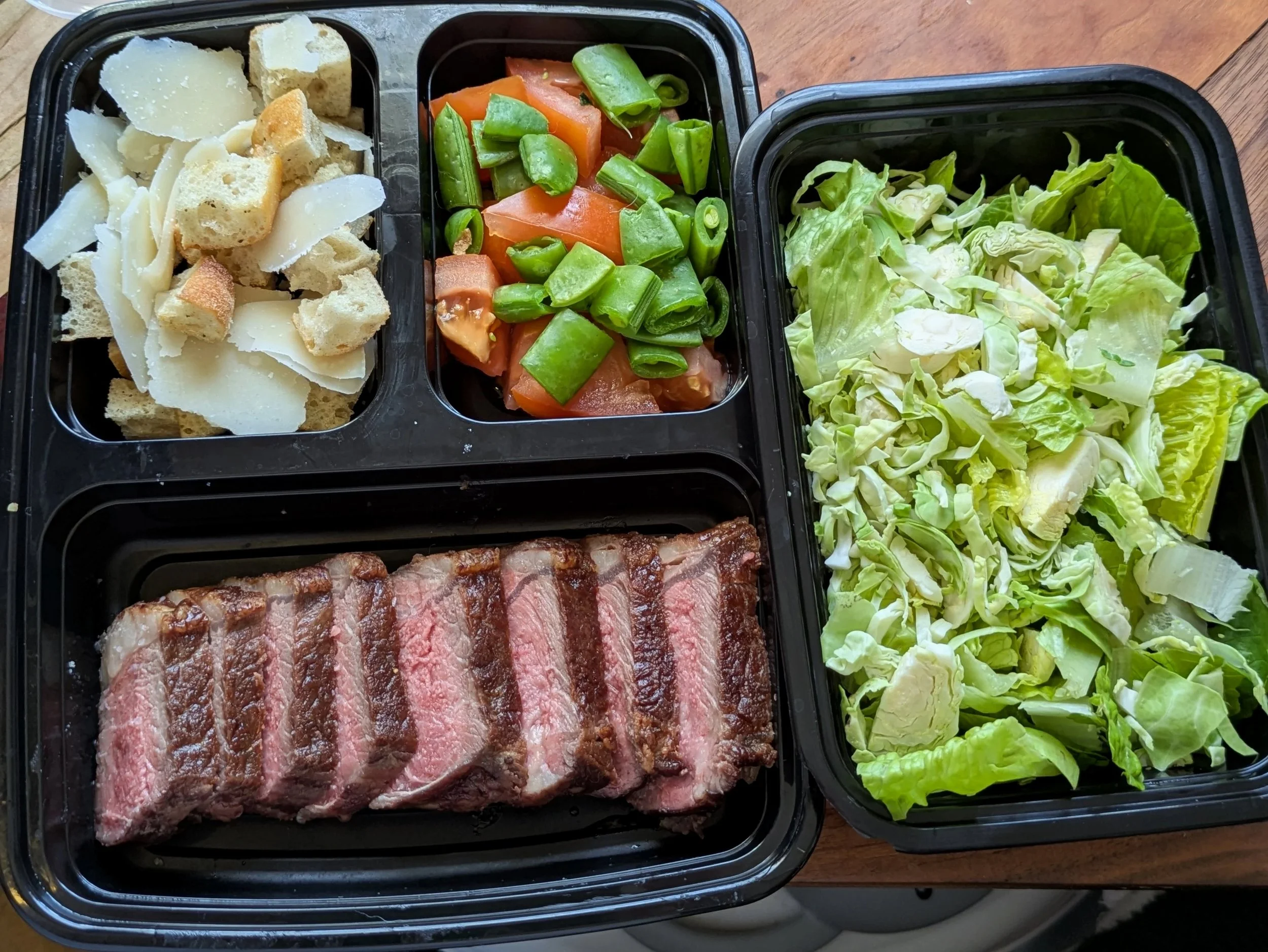 A meal tray with sliced medium rare ny strip steak steak, a romain and shaved brussel sprout salad, a sugar snap pea and tomato mix, and croutons with parmesan cheese.