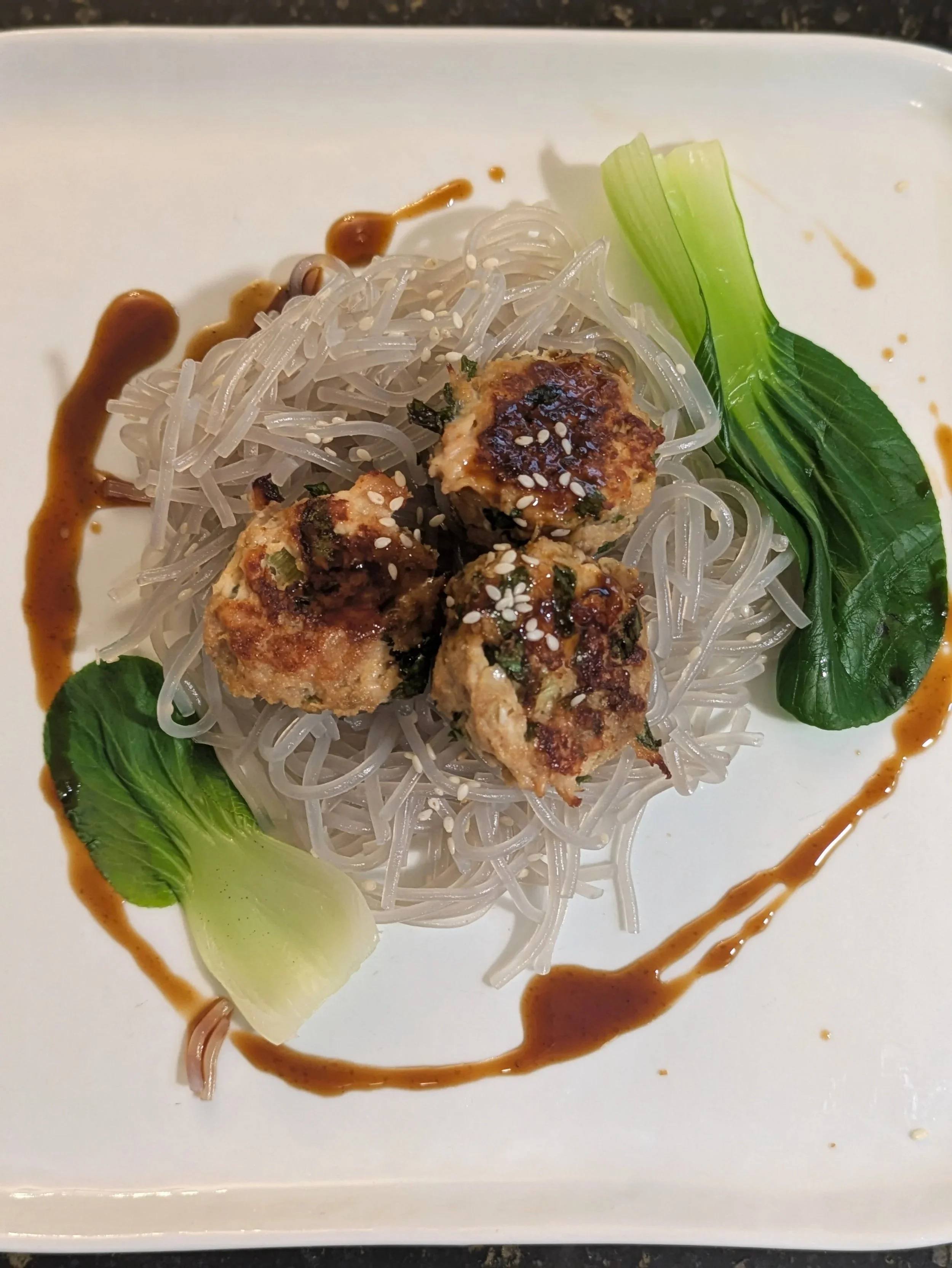 Plate of Asian cuisine featuring three ginger chicken meatballs next to rice atop sweet potato noodles, garnished with sesame seeds, green leafy bok choy, and drizzled with soy glaze.