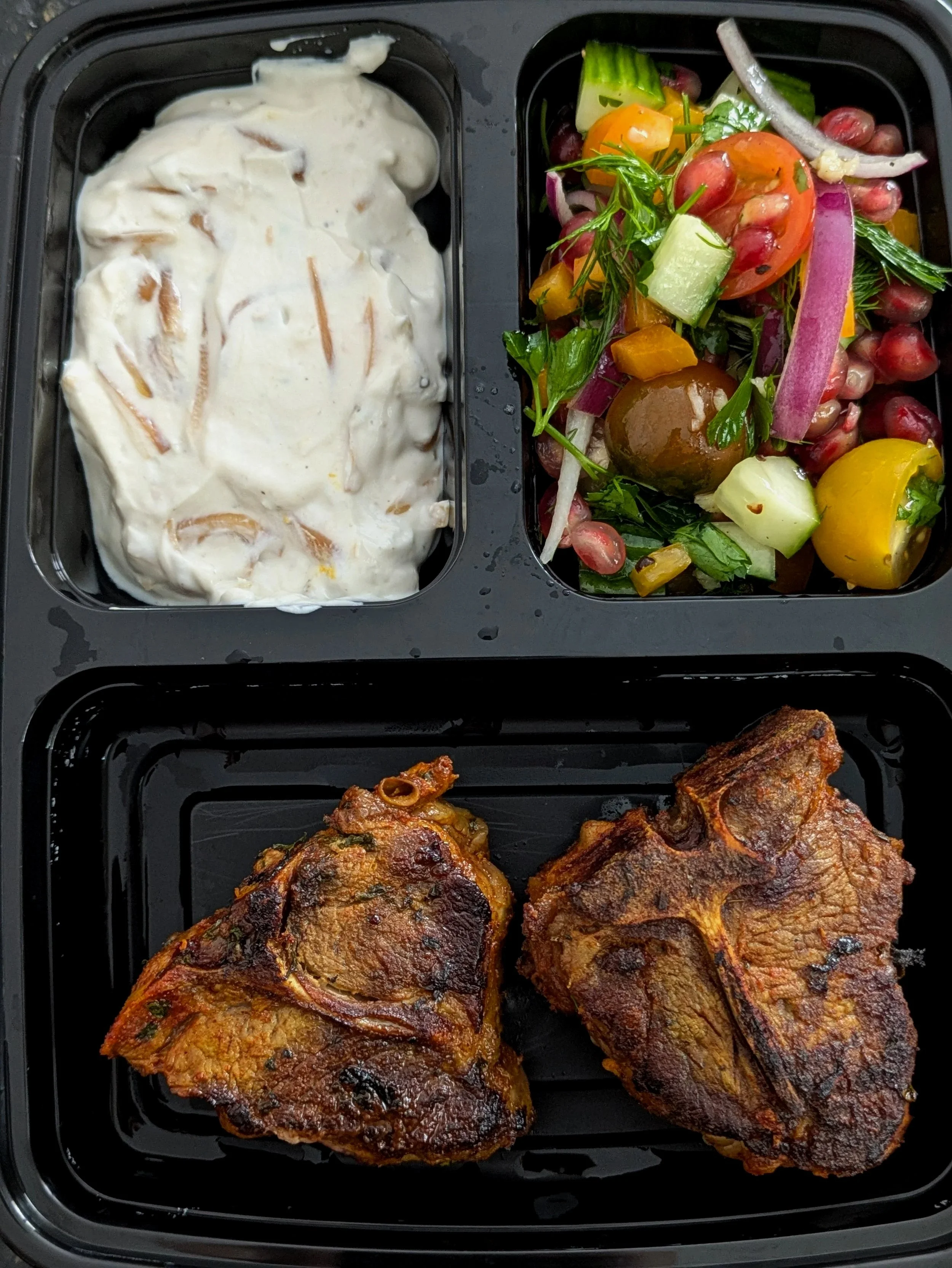 A black compartment meal tray with three sections containing fried chicken, a mixed vegetable salad, and a serving of creamy coleslaw.