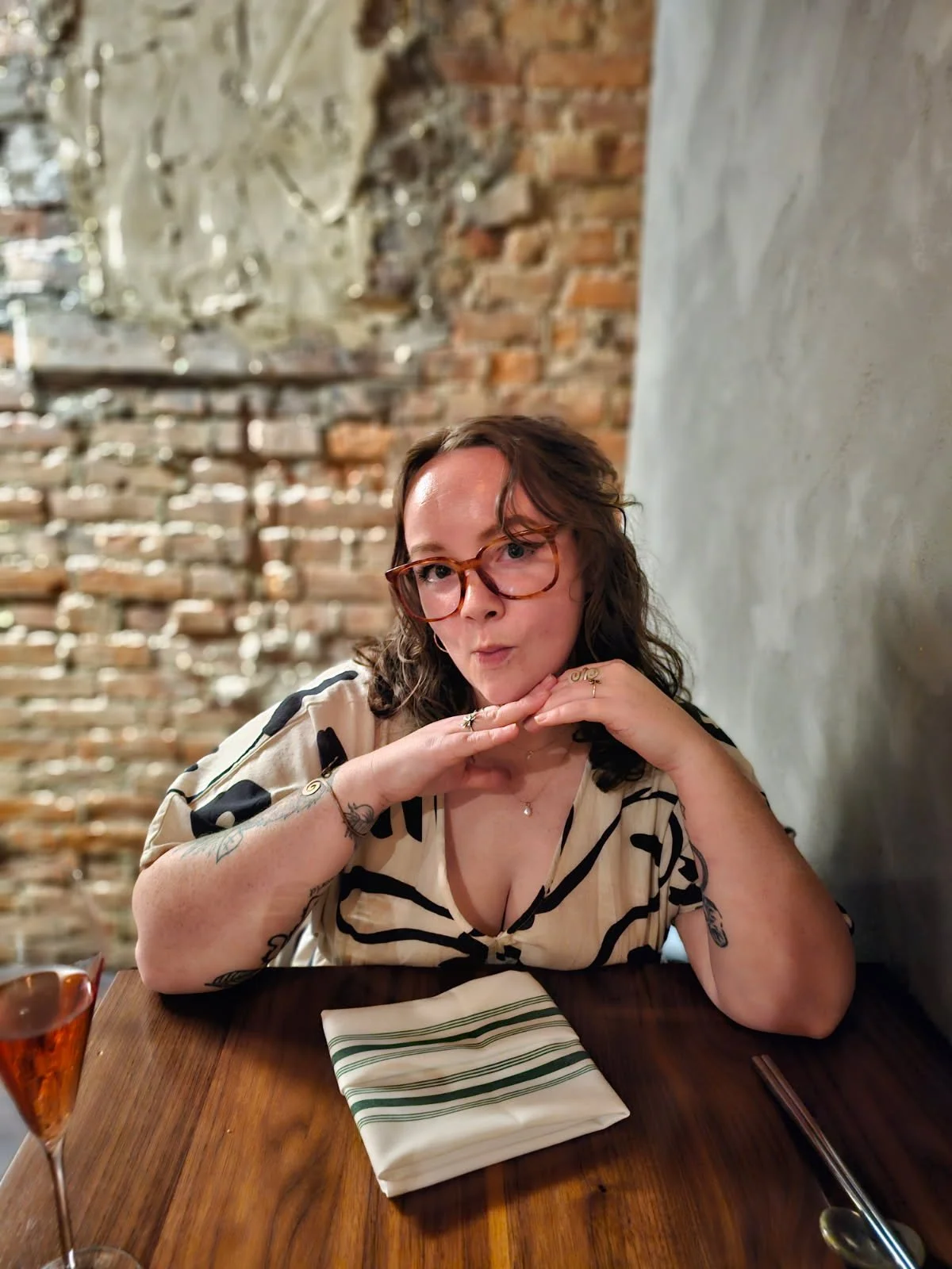 A woman with brown, curly hair and glasses posing with her hands near her face at a restaurant table with a glass of rosé wine and a striped napkin.