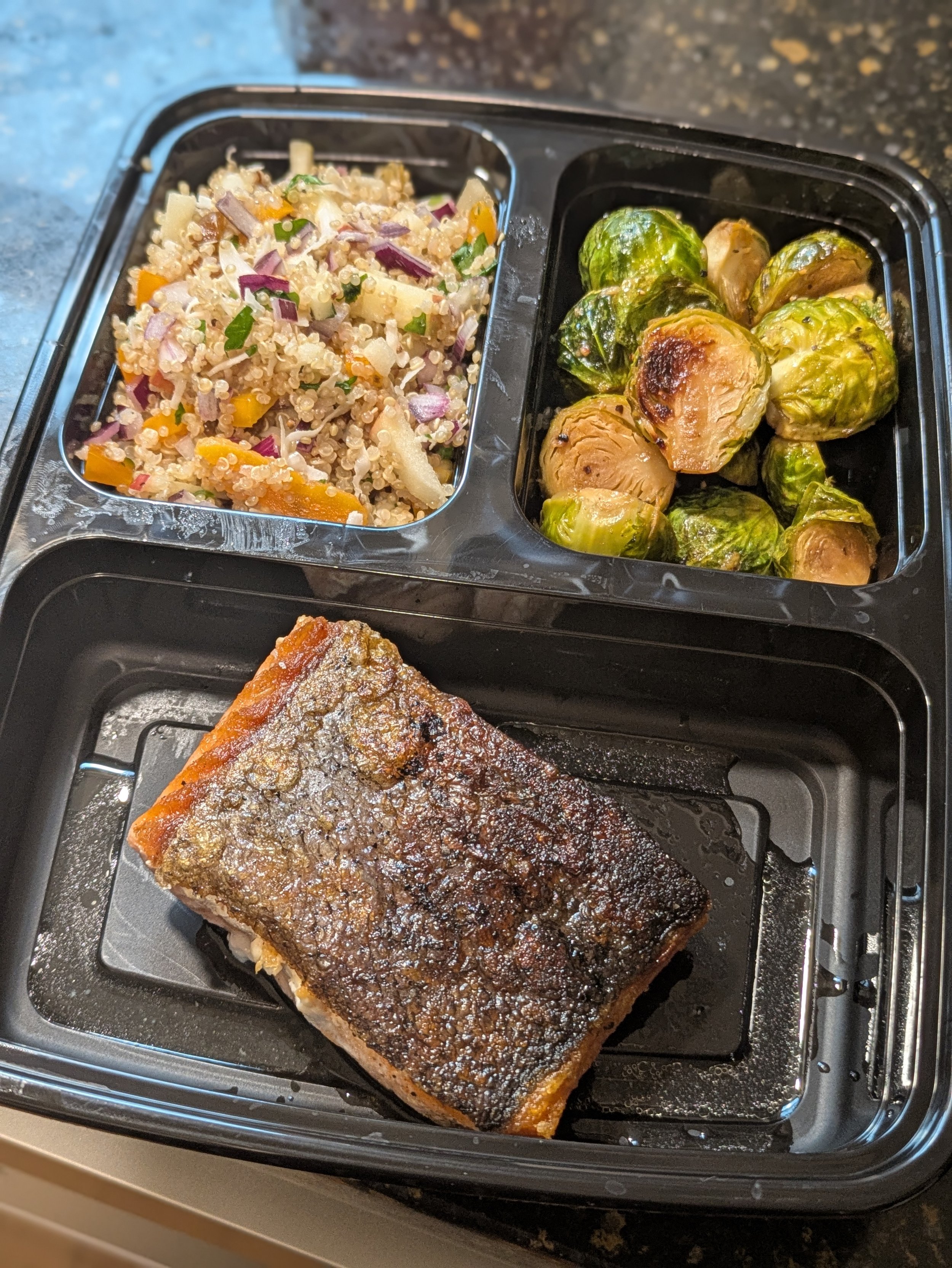 A black divided tray with cooked salmon at the bottom, Brussels sprouts and a quinoa salad with vegetables at the top.