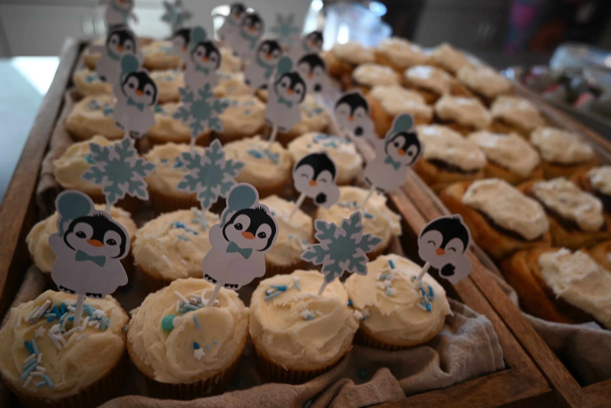 Vanilla bean Cupcakes decorated with blue penguin-themed toppers and snowflake decorations on a wooden display at a baby shower