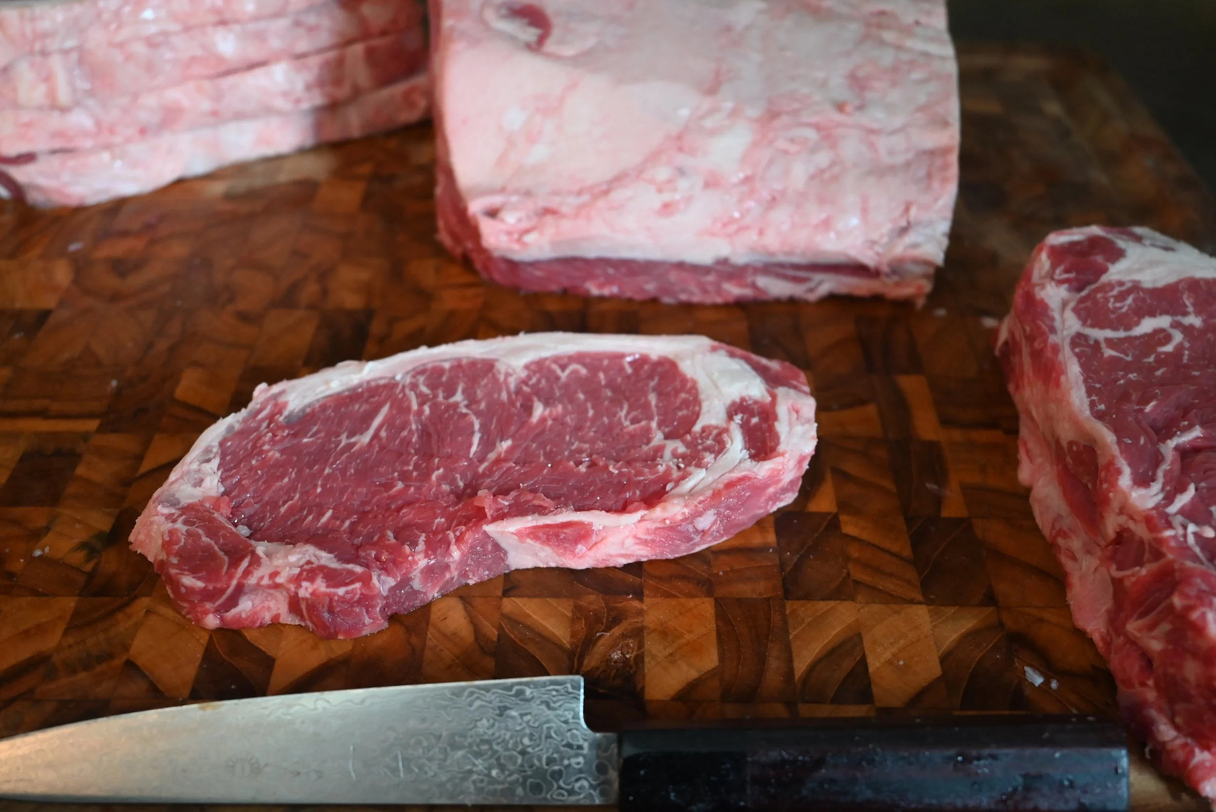 New york strip loin being portioned into Several steaks  on a wooden cutting board with a sharp Japanese petty knife at the bottom left.