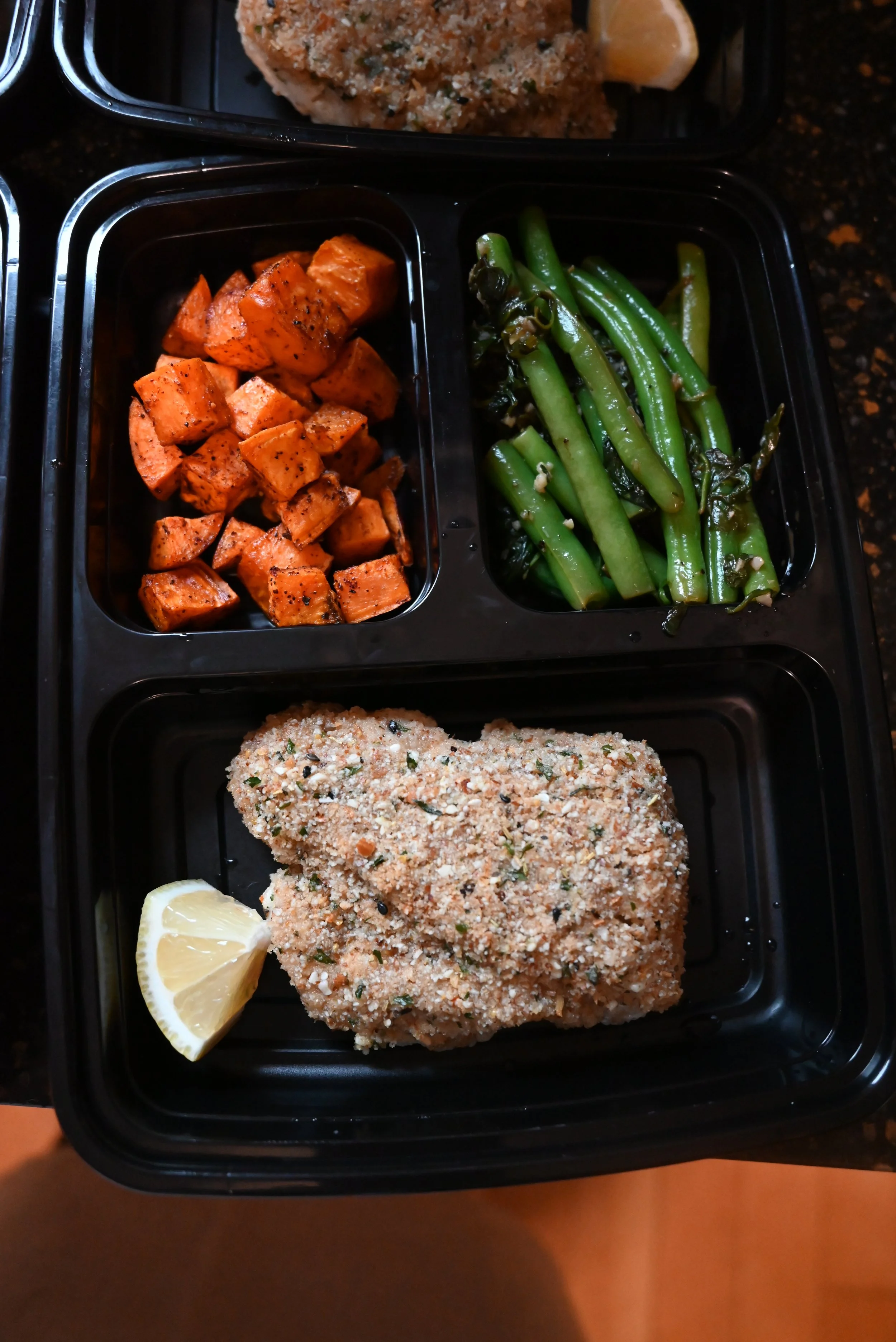 A black divided meal tray with roasted sweet potatoes, sautéed green beans, and a breaded baked fish fillet served with a lemon wedge.