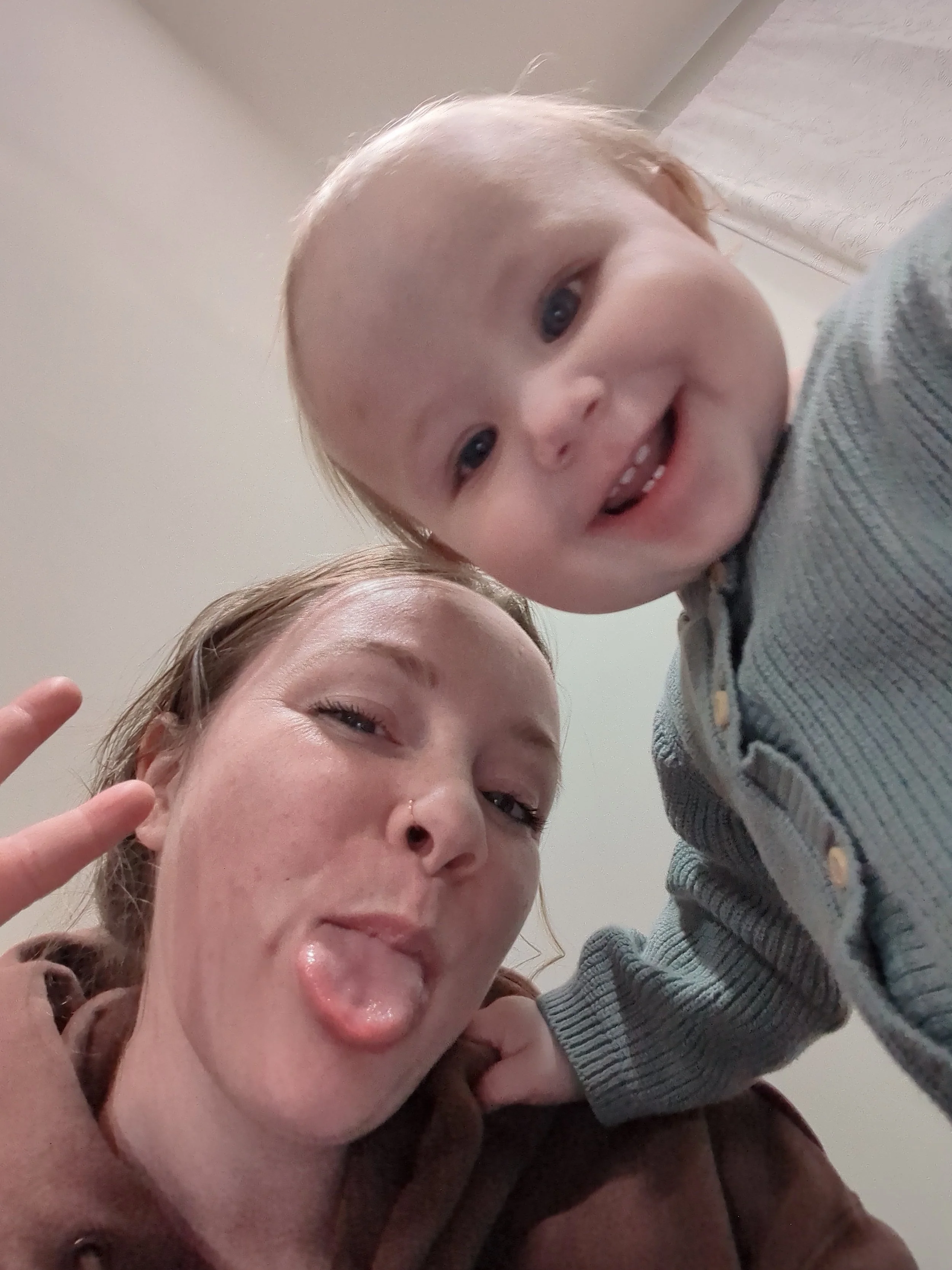 A young woman with red hair and a nose piercing making a funny face with her tongue out and the woman making a peace sign, while a smiling baby with blonde hair and a gray jacket leans in from above.