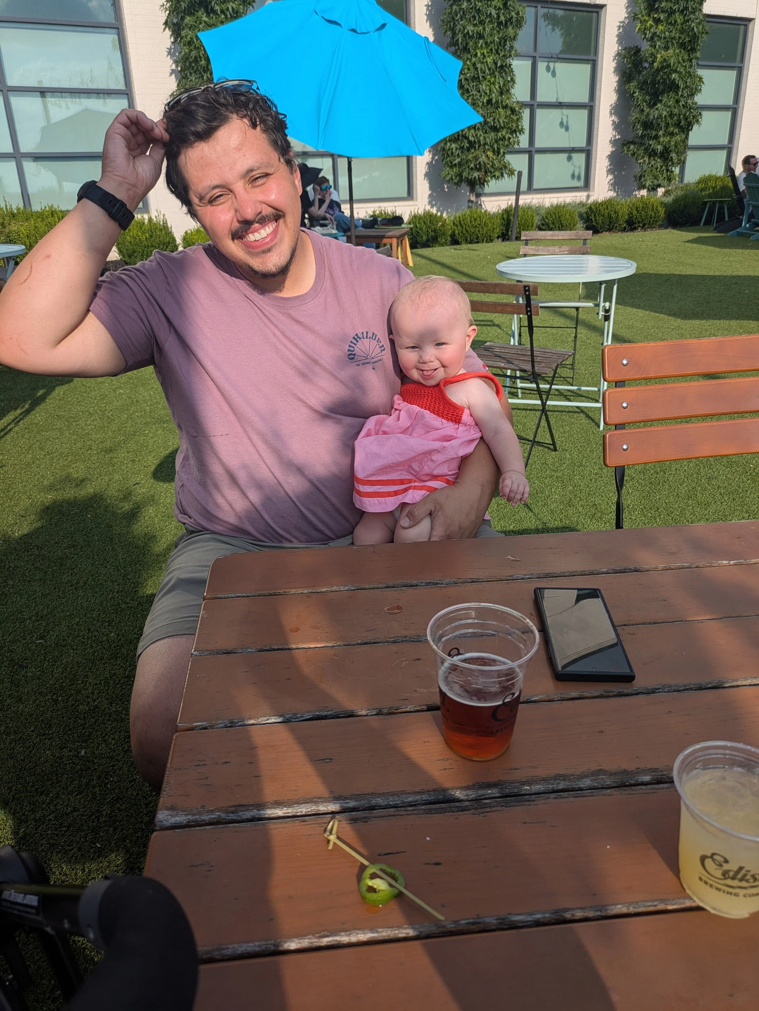 A man with a beard and short dark curly hair sitting outdoors at a wooden table, smiling, holding a baby girl in a pink dress. The baby is also smiling and sitting on his lap. There are drinks on the table, a smartphone, and a green jalapeño pepper with a yellow toothpick. The background shows green grass, outdoor furniture, a building with large windows, and a blue umbrella.