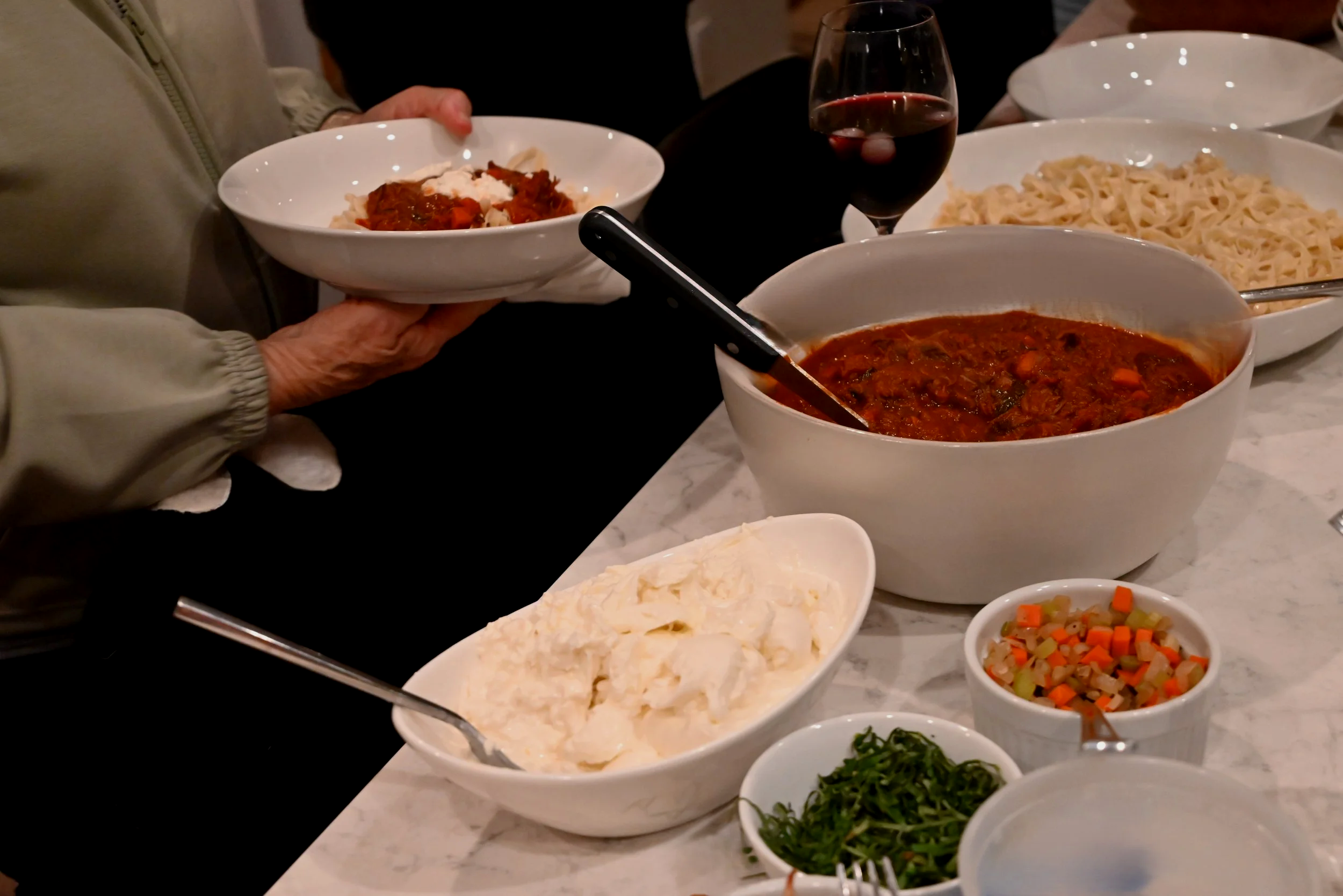 A table with bowls of pasta, soup, mashed potatoes, mixed vegetables, chopped greens, and a glass of red wine. A person is holding a bowl of pasta.