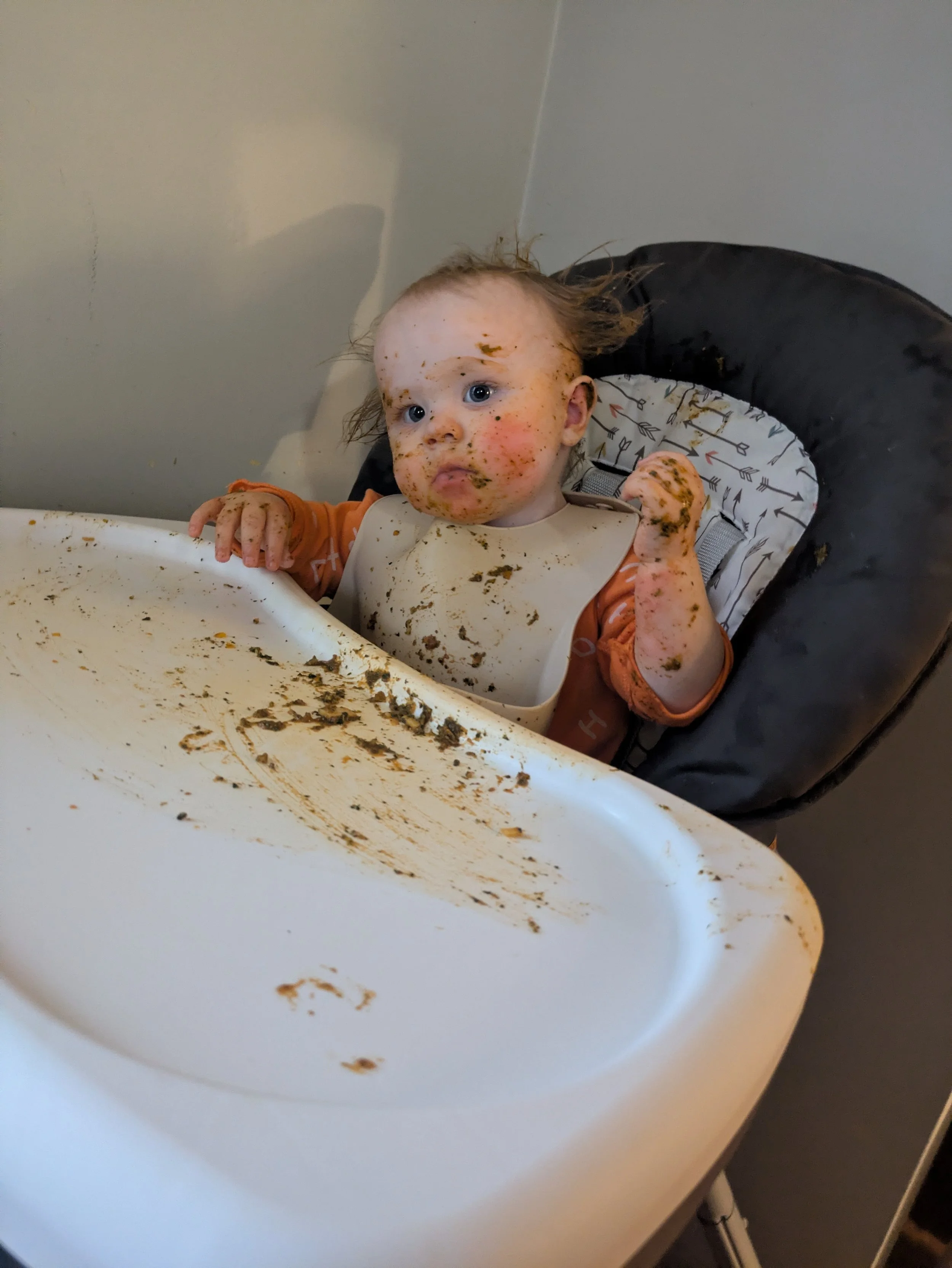 A young child with messy face and hands covered in food, sitting in a high chair with a dirty tray, looking up with a curious expression.