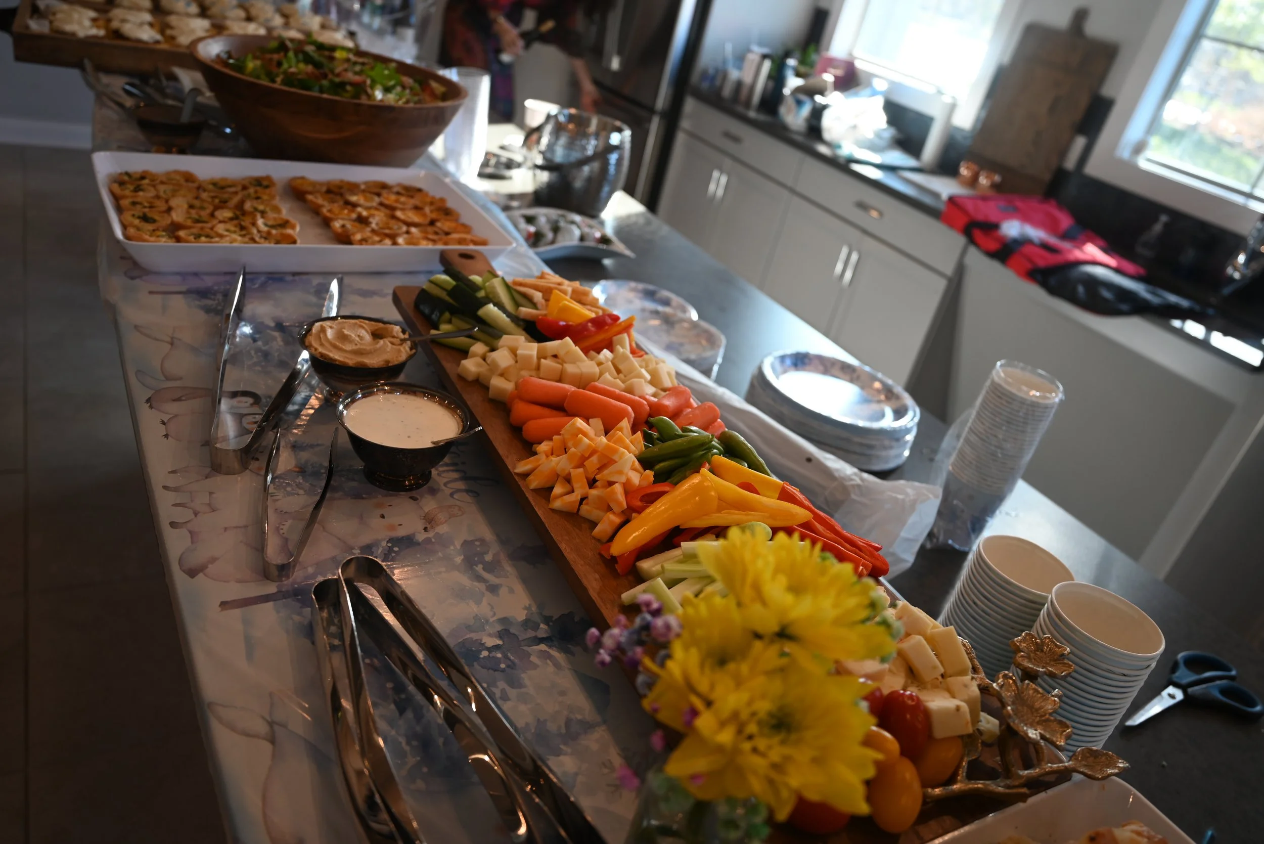 Buffet table with cheese, vegetables, salads, and appetizers in a kitchen with cabinets and a window in the background.