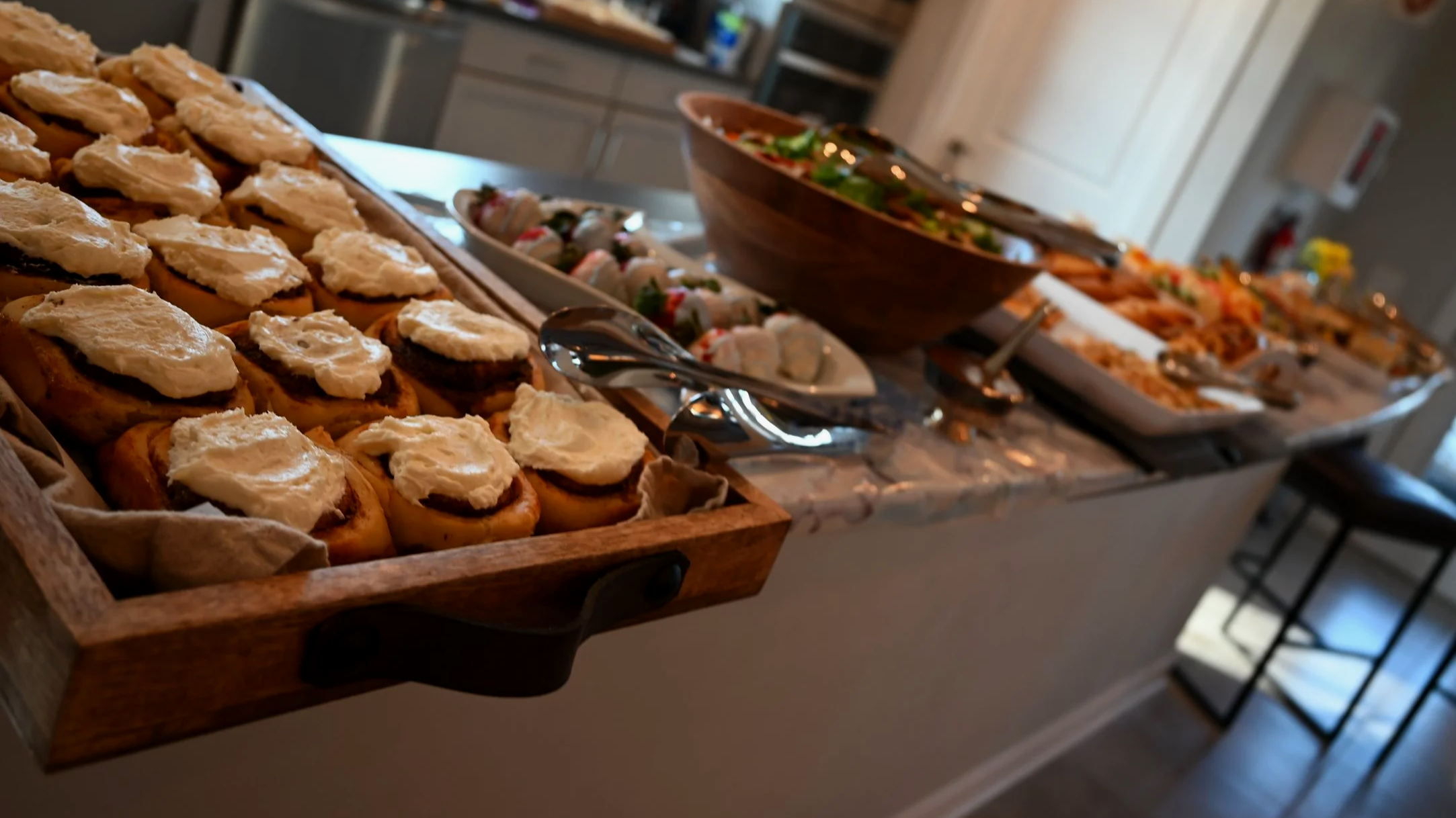 Buffet table with various appetizers including sliders with cheese and meat, sushi rolls, salads, and other finger foods, set in a casual indoor setting.