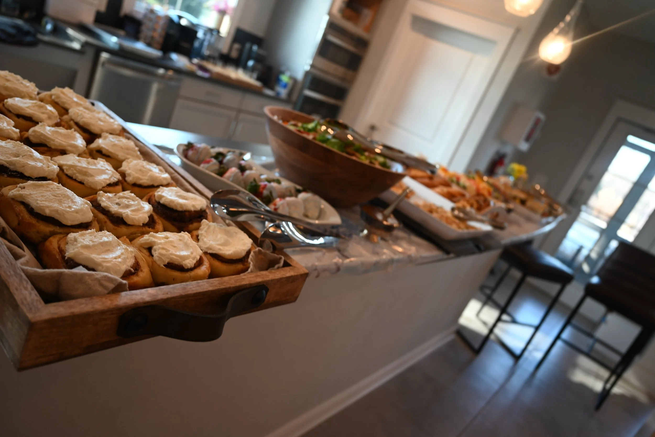 Buffet table with various dishes including tea sandwiches with white topping, vegetable platter, and other finger foods in a bright, modern kitchen or dining area.