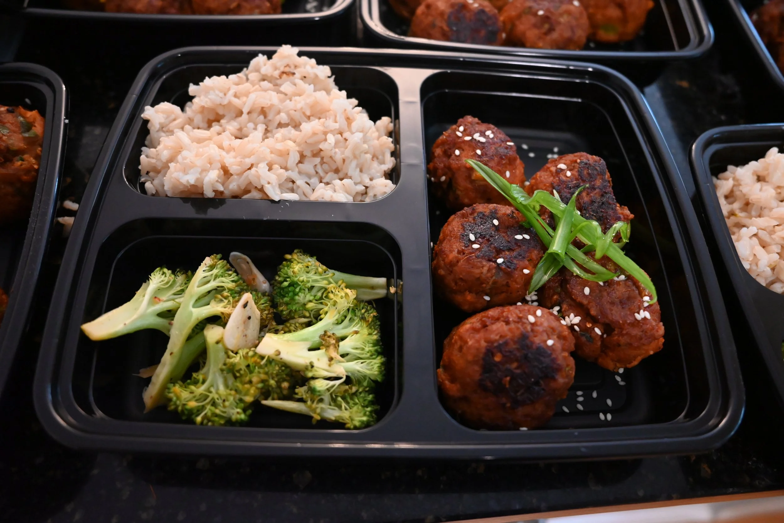 A black meal tray with three compartments containing rice, broccoli, and meatballs garnished with green onions and sesame seeds.