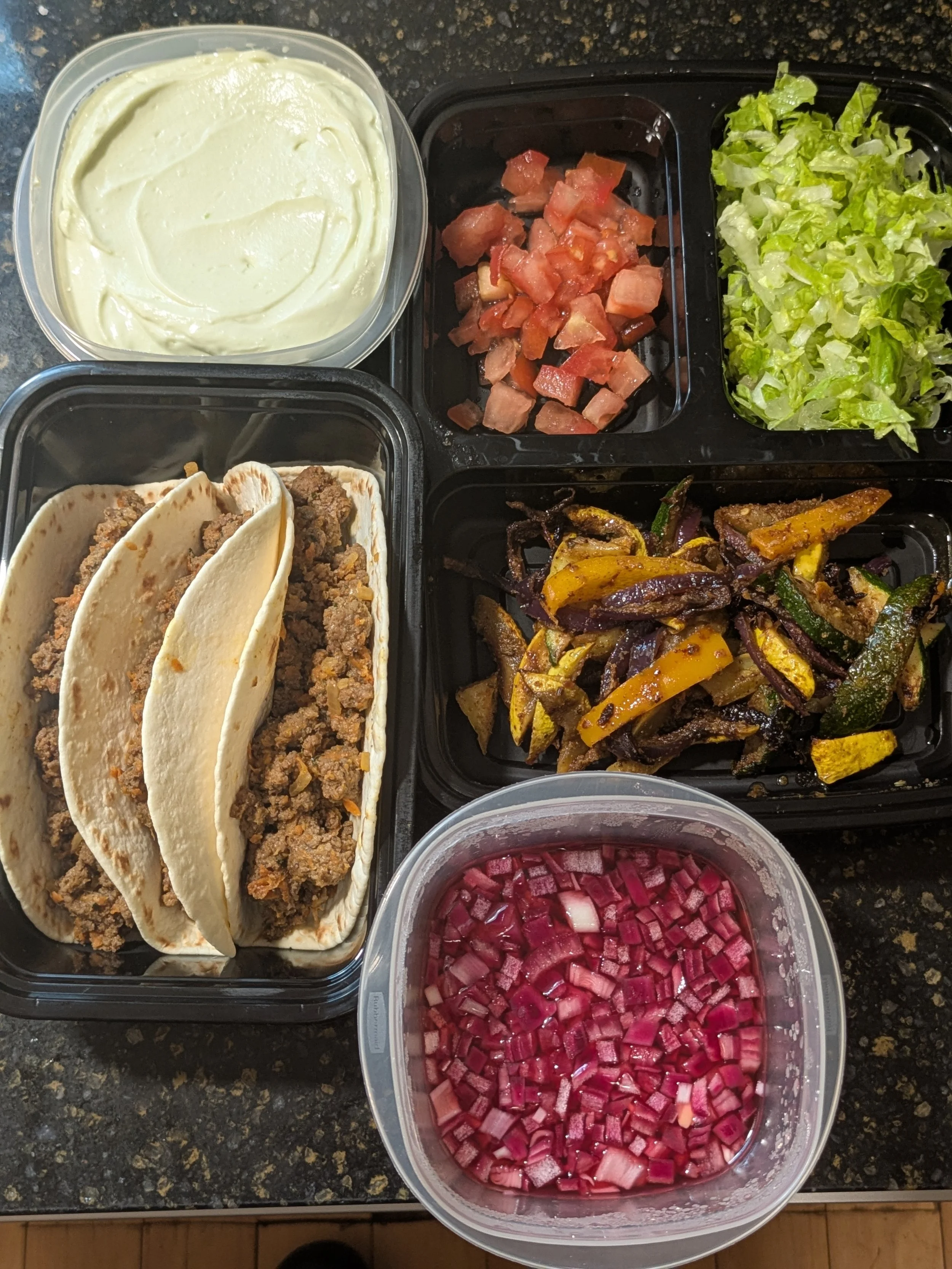 A meal tray with beef tacos, yellow rice, chopped tomatoes, chopped red onions, shredded lettuce, seasoned grilled vegetables, and sour cream.