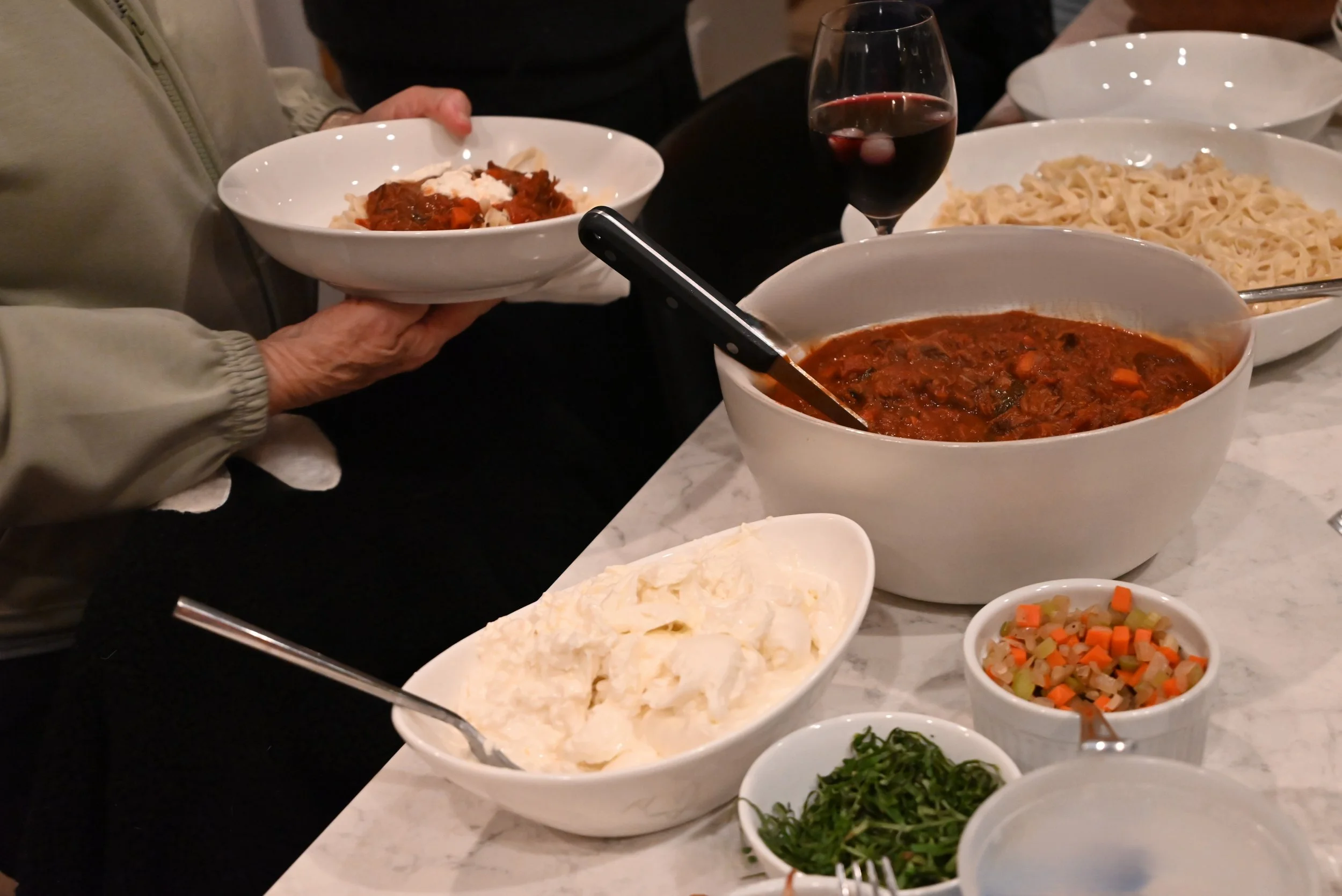 A table with various dishes including a large bowl of beef stew, bowls of mashed potatoes, chopped vegetables, fresh herbs, and a glass of red wine, with people serving themselves
