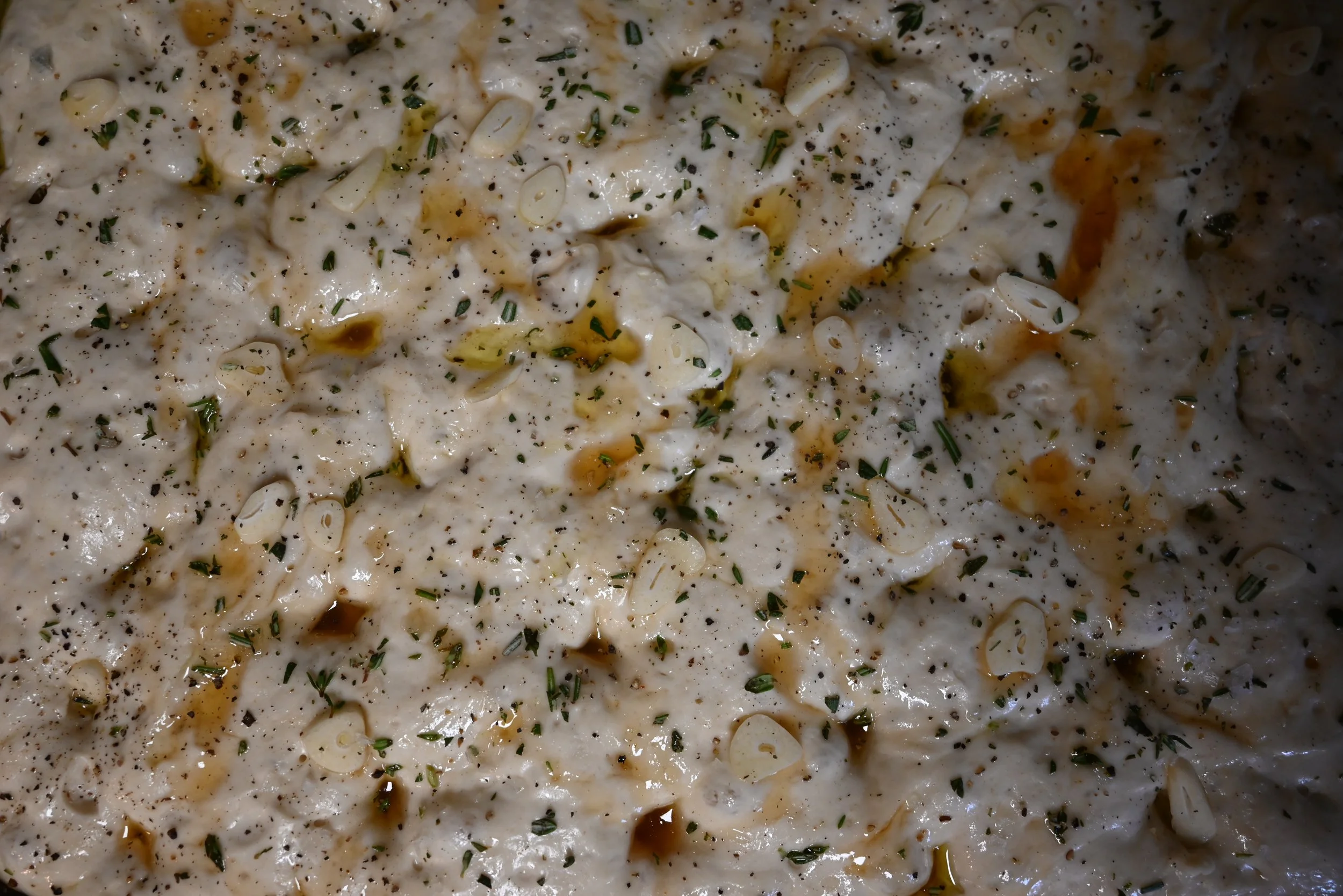 Close-up of an un-baked dimpled focaccia garnished with herbs, black pepper, sliced green onions, and drizzled with oil.