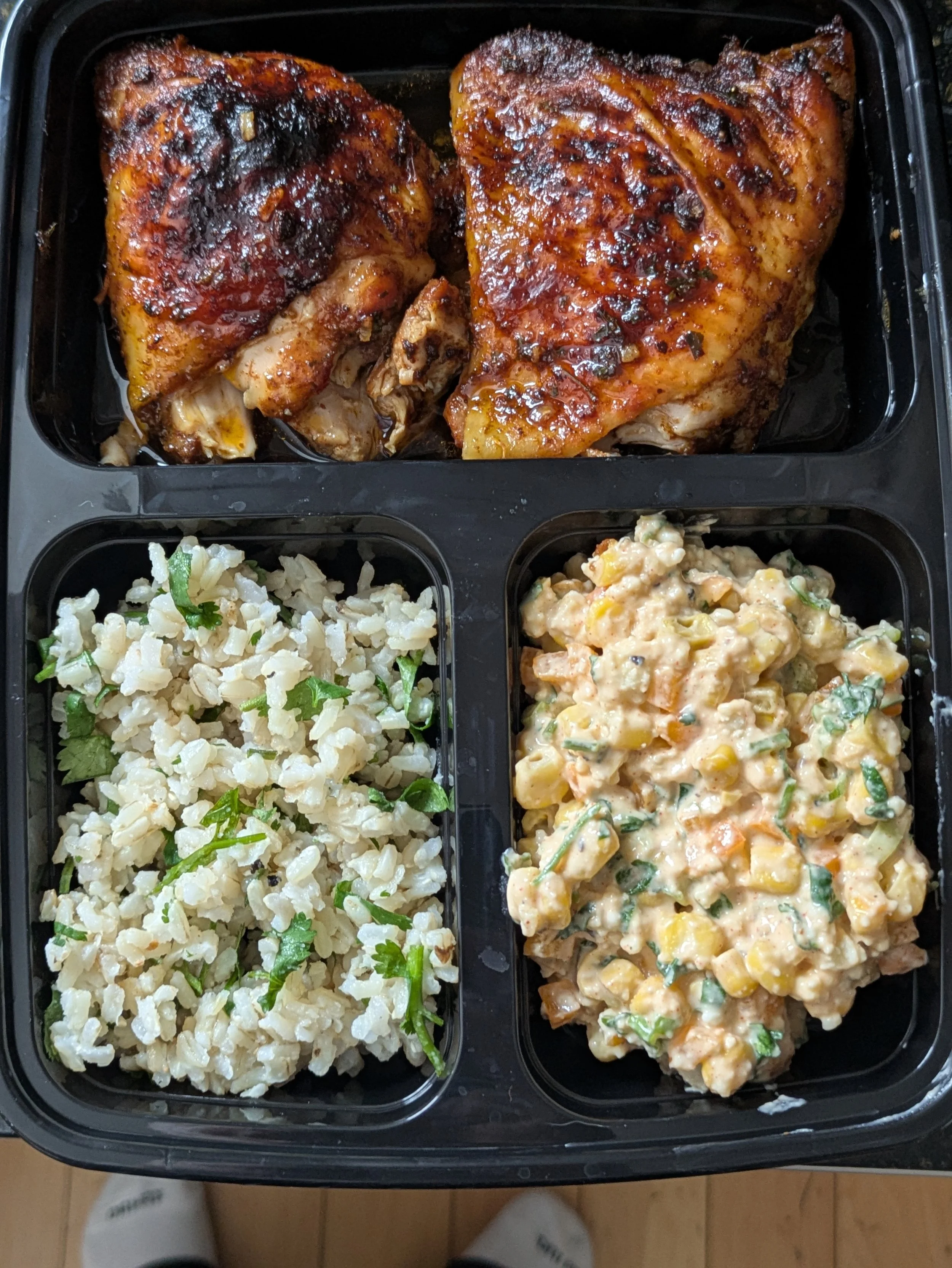 Meal tray with two grilled chicken thighs topped with barbecue sauce in the top compartment, a serving of rice salad with herbs in the bottom left, and a creamy chicken salad with vegetables in the bottom right.