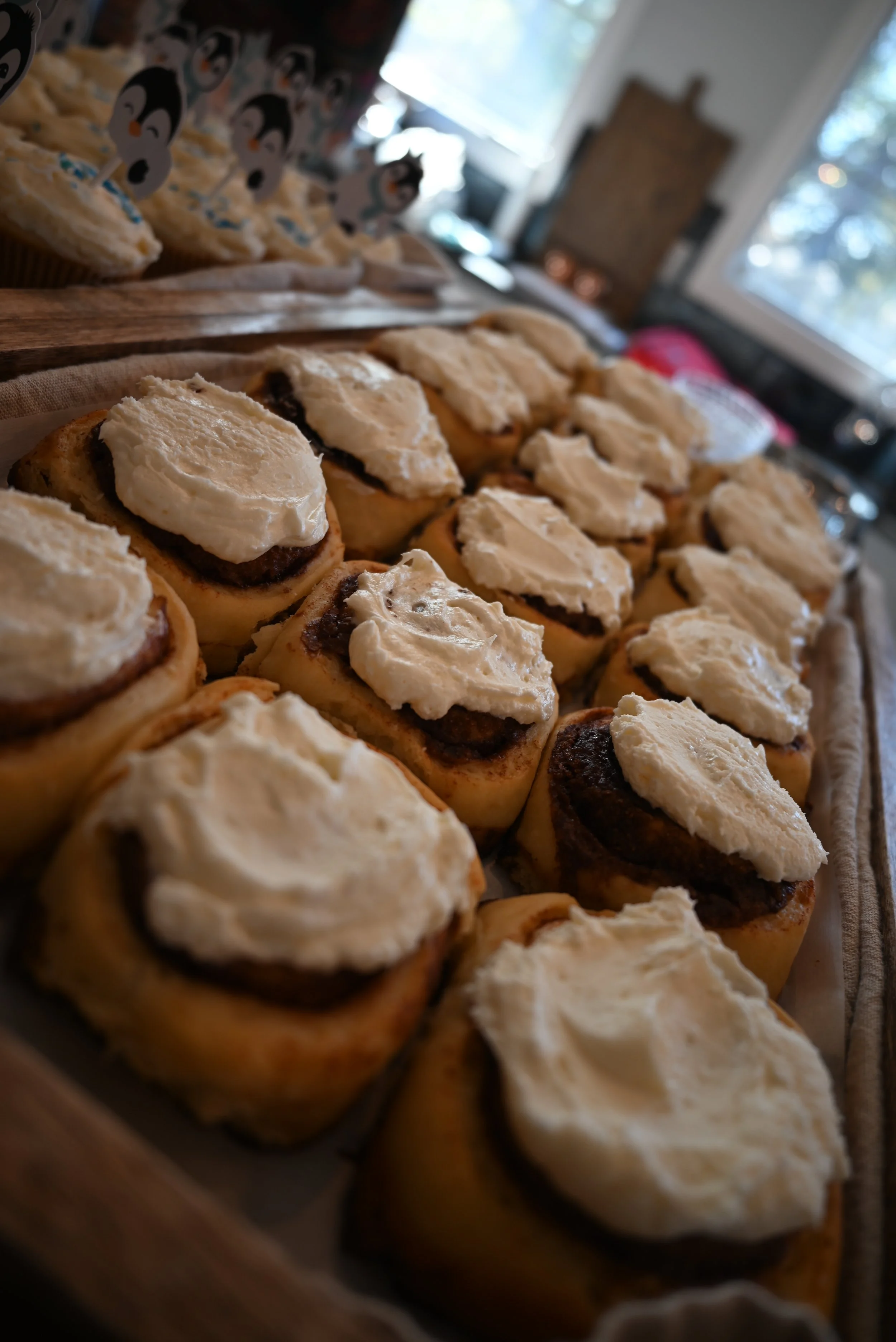 A tray of fresh baked cinnamon rolls topped with cream cheese frosting, with cupcakes decorated with penguin toppers visible in the background.