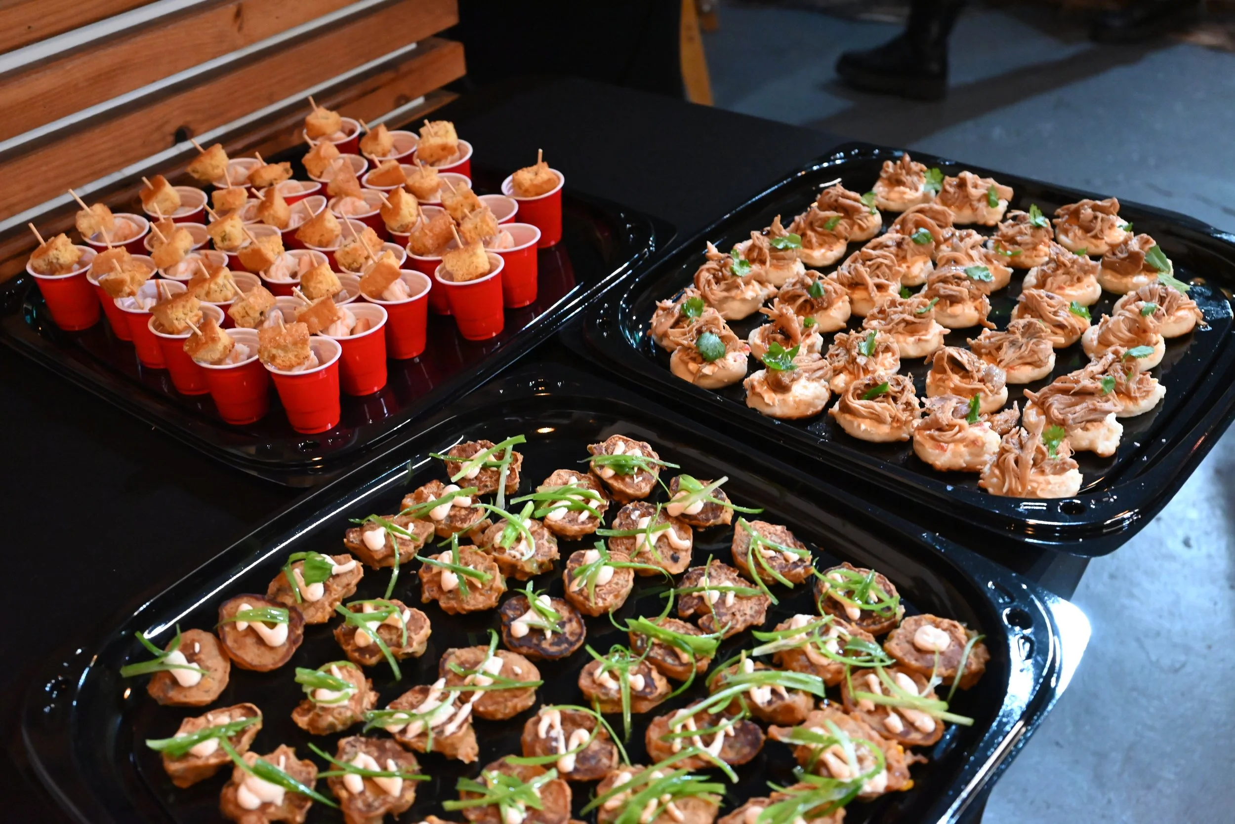 Tray of bite-sized appetizers, including small cups with skewered fried food, topped miniature sandwiches with shredded meat and green herbs, and fried pieces garnished with green onion and white sauce.