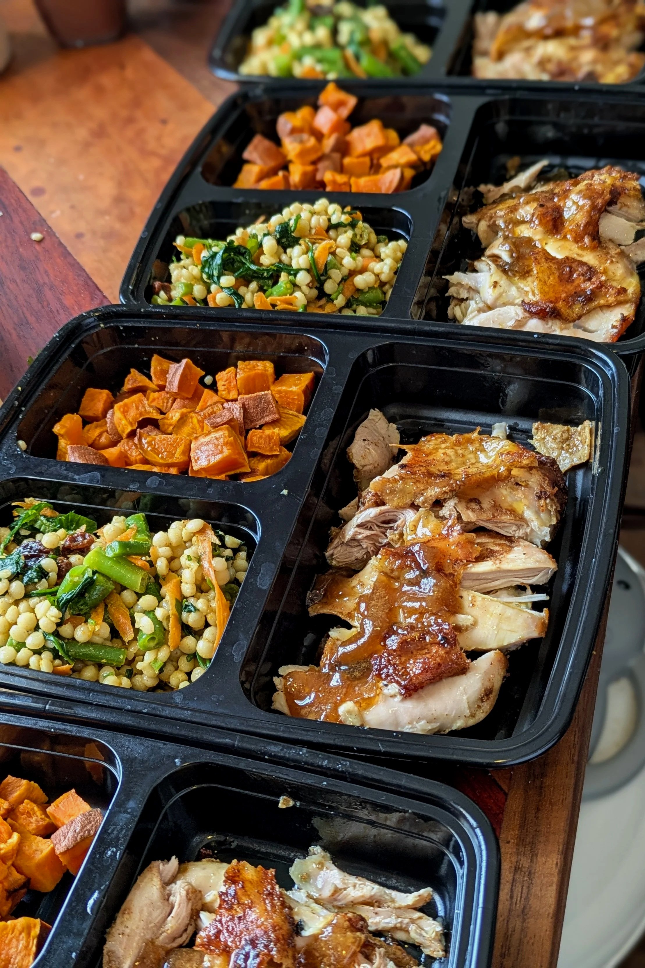 Black meal prep containers filled with cooked chicken, roasted sweet potatoes, and mixed salads on a wooden surface.