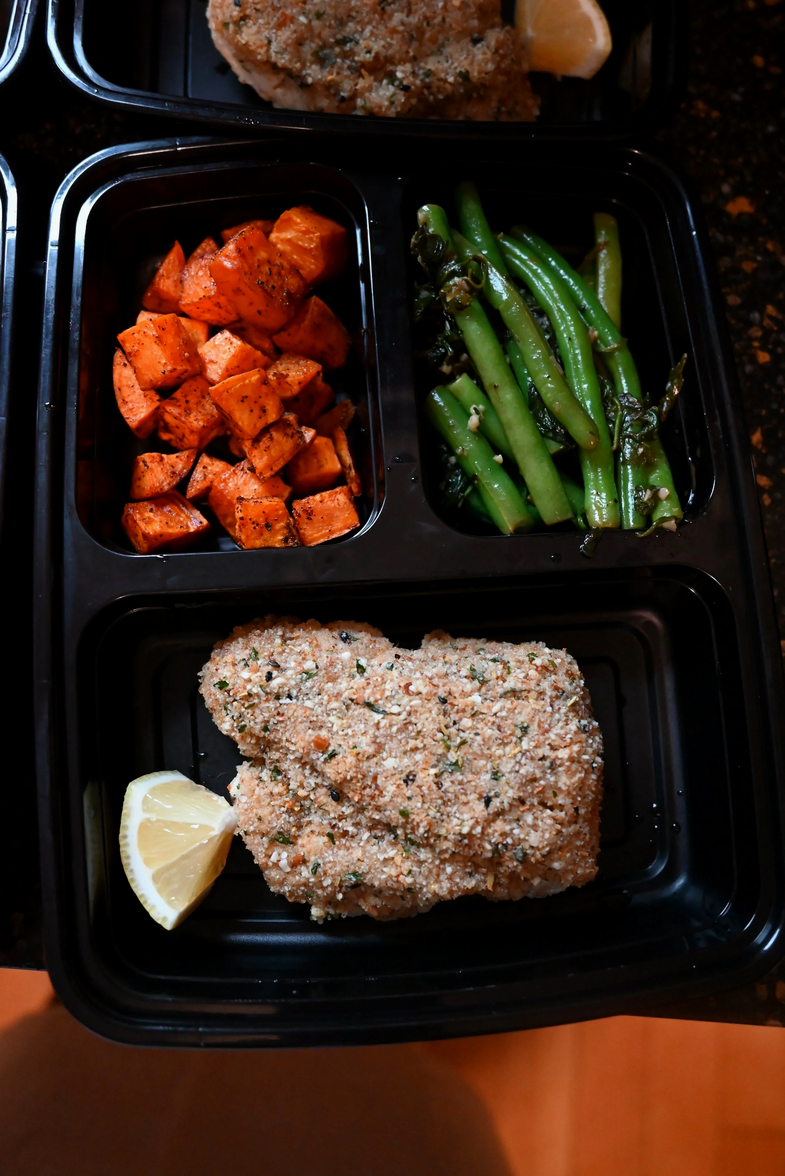Black tray with three compartments containing roasted sweet potatoes, sautéed green beans, and a breaded piece of fish with a lemon wedge.