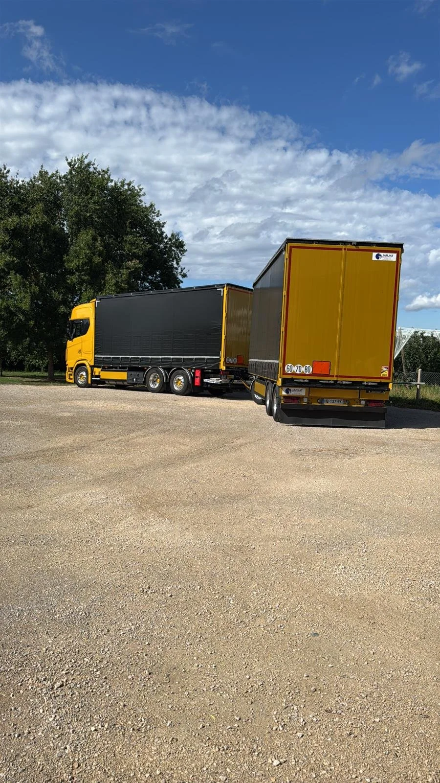 Un camion remorque jaune avec des bâches noires, garés sur un sol de gravier, avec un ciel partiellement nuageux et des arbres en arrière-plan.