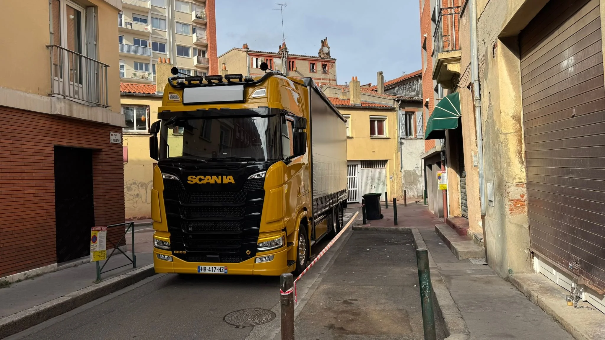 Un camion jaune de la marque Scania stationné dans une ruelle urbaine, entouré de bâtiments résidentiels avec des façades en brique et en plâtre, sous un ciel clair.