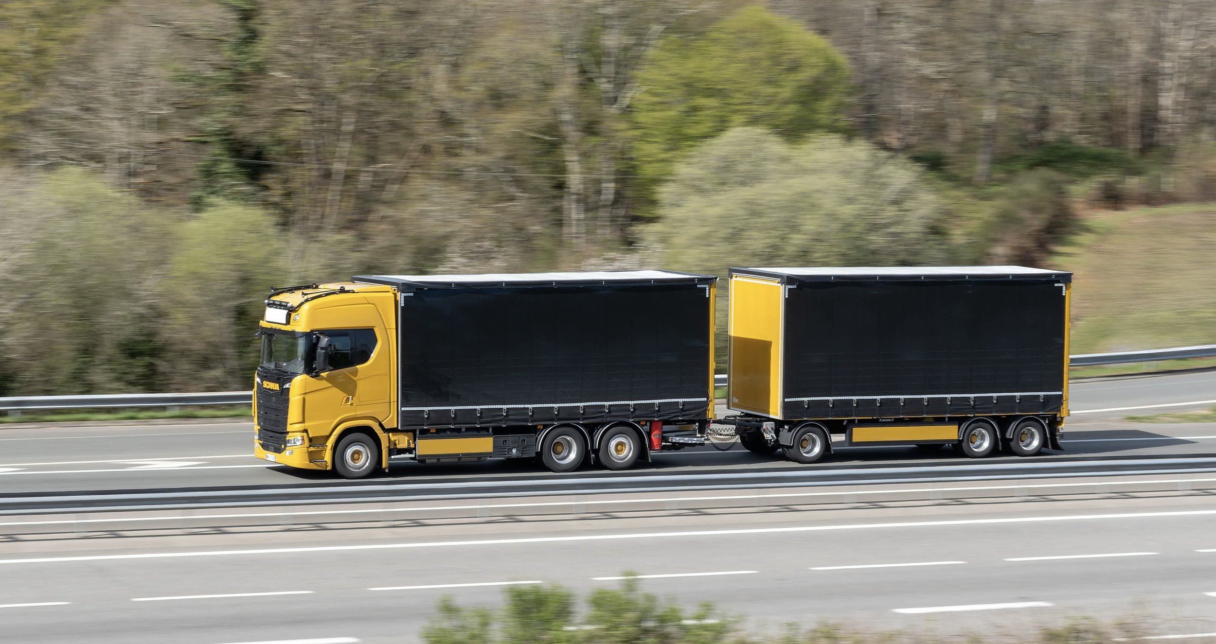 Camion jaune avec remorque noire roulant sur une autoroute, avec un arrière-plan de collines et d'arbres.